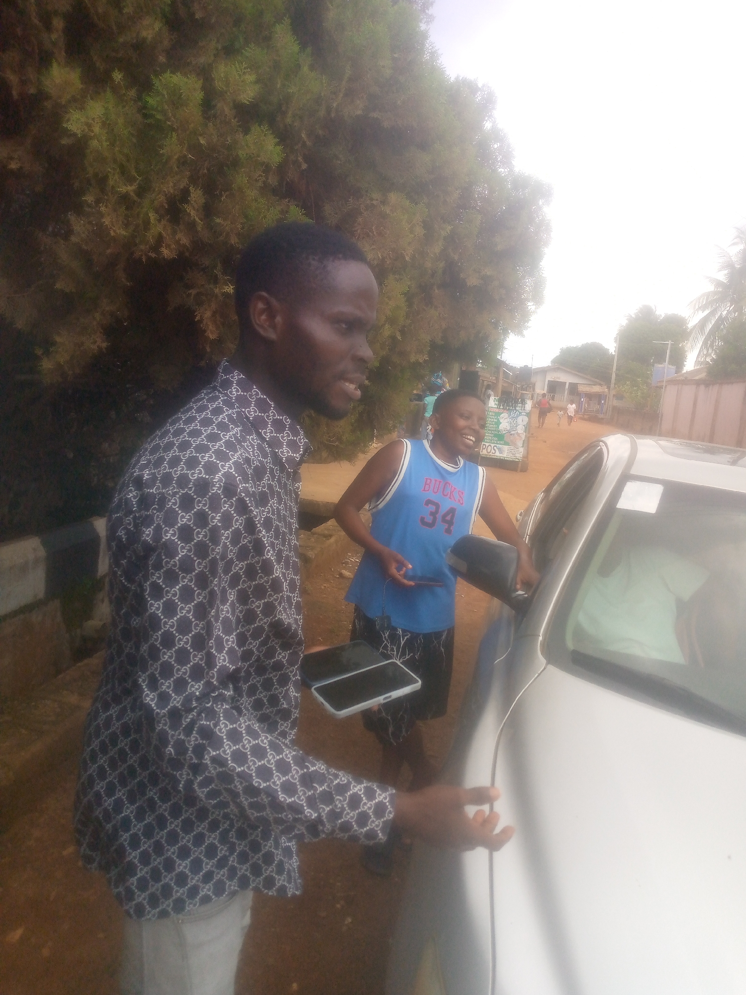 Two men are outside, next to a silver car. The man on the left is wearing a dark blue and white patterned shirt and light-colored pants, holding a smartphone. The man on the right is wearing a blue basketball jersey with "BUCKS" and the number "34" printed on it, holding a mobile phone, smiling, and leaning on the car. The background reveals a dirt road with buildings and people in the distance. Trees and vegetation surround the scene. The setting appears to be a rural or suburban area. The location is Unknown, Unknown.