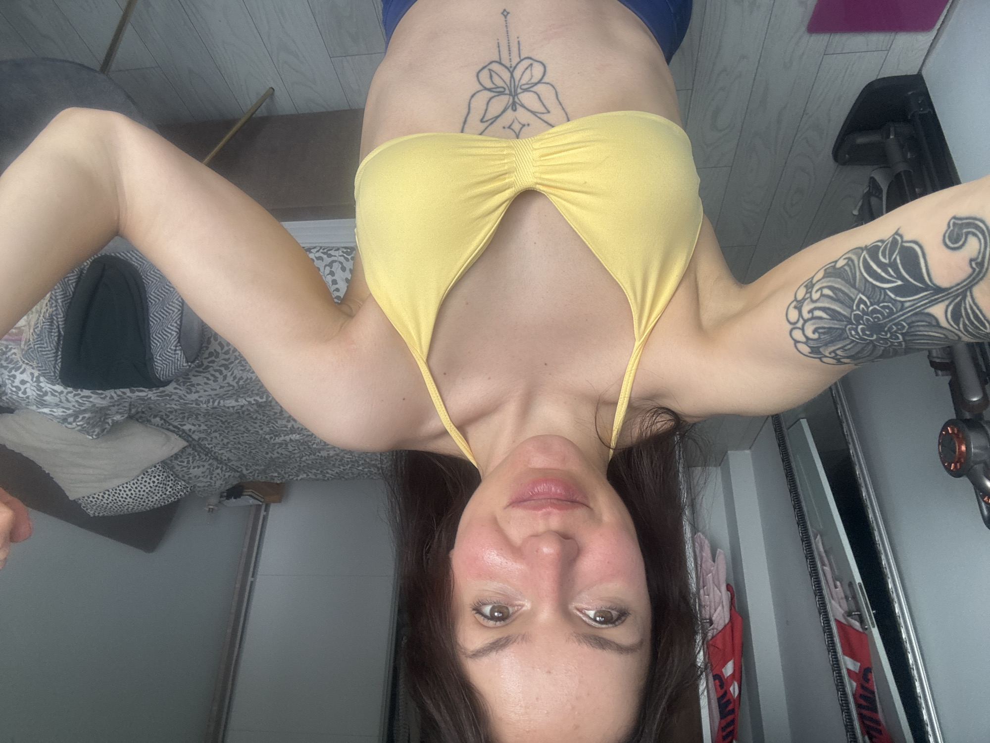 A woman is posing for a selfie, holding the camera above her and angled down towards her face. She is wearing a yellow bikini top and blue bottoms. She has dark hair and fair skin, with a tattoo on her stomach and another on her upper left arm.
The background includes parts of a room: a bed with patterned bedding, a gray wall, a mirror revealing items within a closet, a vacuum cleaner, and a chair. The image is rotated. The location in the city of Burgas, Bulgaria, is not discernible.