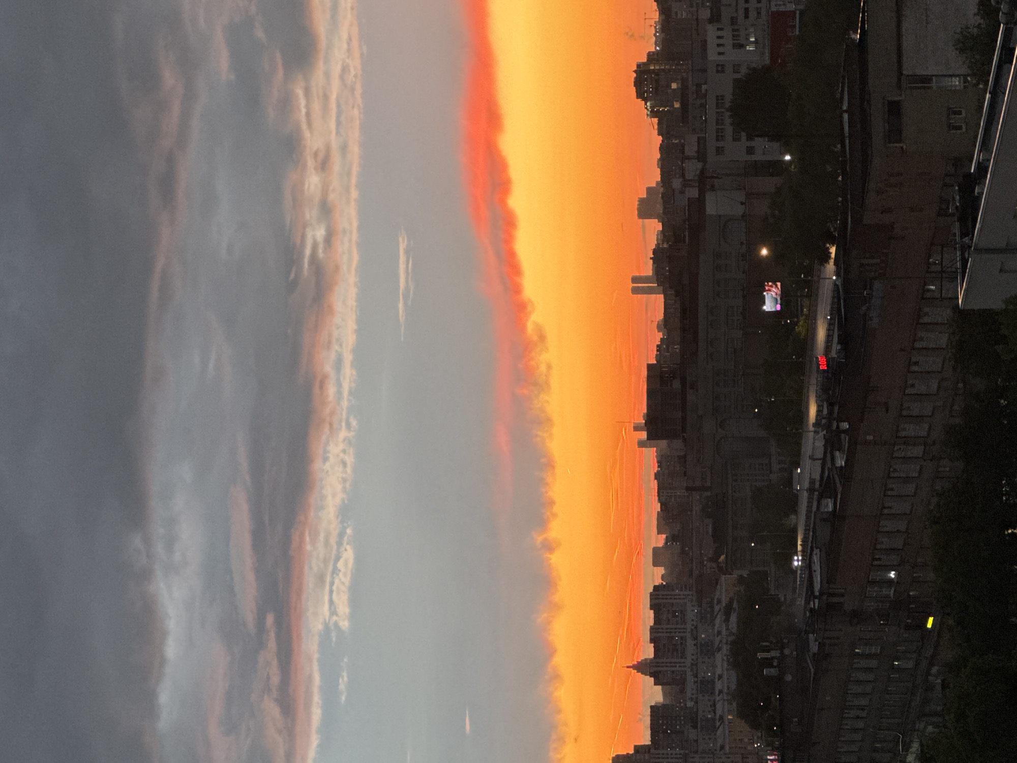 An aerial, eye-level, medium shot captures a city skyline at dusk. The sky is a vibrant gradient of fiery orange and yellow at the horizon, transitioning to pink and grey clouds above. Silhouetted against the bright sky are numerous buildings of varying heights, characteristic of a dense urban environment. The architecture suggests a mix of older, mid-rise structures and taller modern buildings, including what appear to be two distinctive twin towers in the distance. The foreground shows the tops of several darker, lower-rise buildings, some with visible windows and rooftop details. The overall impression is of a sprawling metropolis bathed in the dramatic light of sunset.
