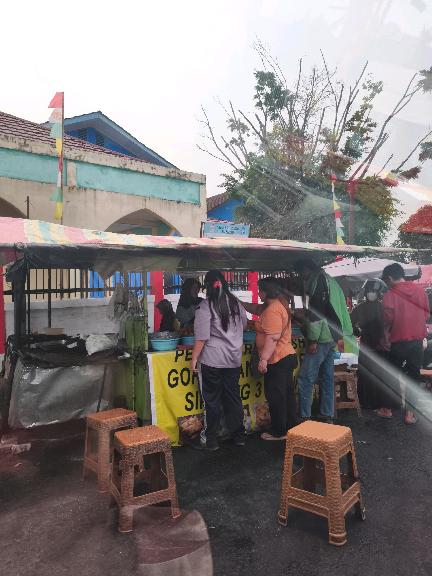 A roadside food stall is depicted in Palembang, Indonesia. A tarp-covered cart serves as the stall, displaying various food items and a yellow banner with Indonesian text that reads "PE GORI SING 3". Several individuals are gathered around the stall, some appearing to be customers and others working. There are three brown plastic stools placed in front of the stall. In the background, a building with a blue roof and turquoise trim is visible, along with trees and a clear sky. The overall scene suggests a typical street food vending operation.