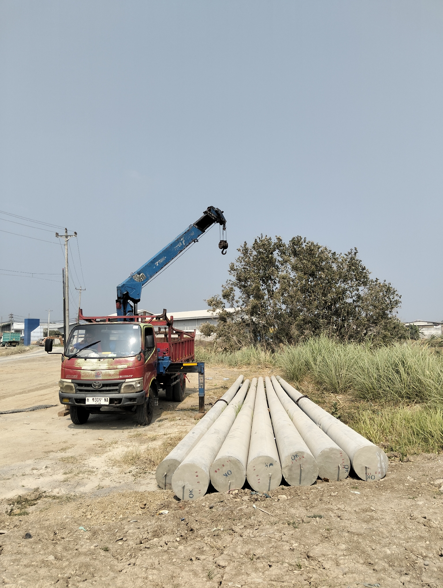 In Semarang, Indonesia, a red truck with a blue crane is parked on a dirt lot. Several large concrete pipes are lying on the ground in front of the truck. The truck has a license plate that reads "H 9309 NA". A person is visible in the driver's seat of the truck. In the background, there is a large tree and some tall grass. There are also some industrial buildings in the distance. The sky is clear and blue.
