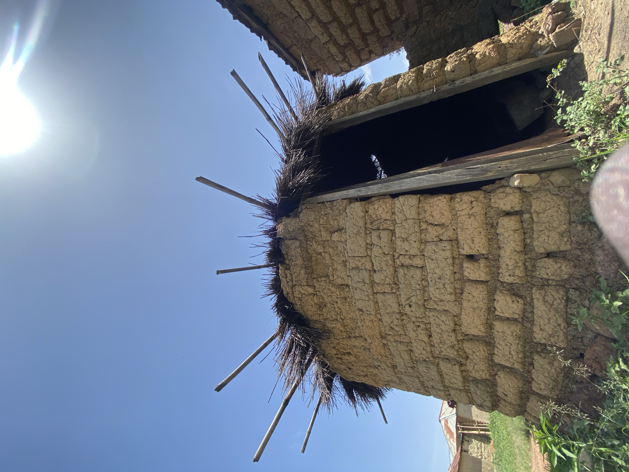 The image depicts a dilapidated mud structure, likely a building or part of a building, under a bright blue sky with the sun visible in the upper left corner. The structure appears to be made of sun-dried mud bricks or adobe and has a thatched roof with protruding wooden supports. A dark opening is visible in the structure, suggestive of a window or doorway. Vegetation, including grasses and small plants, grows near the base of the structure. The image is angled upwards, emphasizing the height and disrepair of the building. There is no evidence in the image of the presence of people. There is no proof that this is in Durimi, Nigeria.
