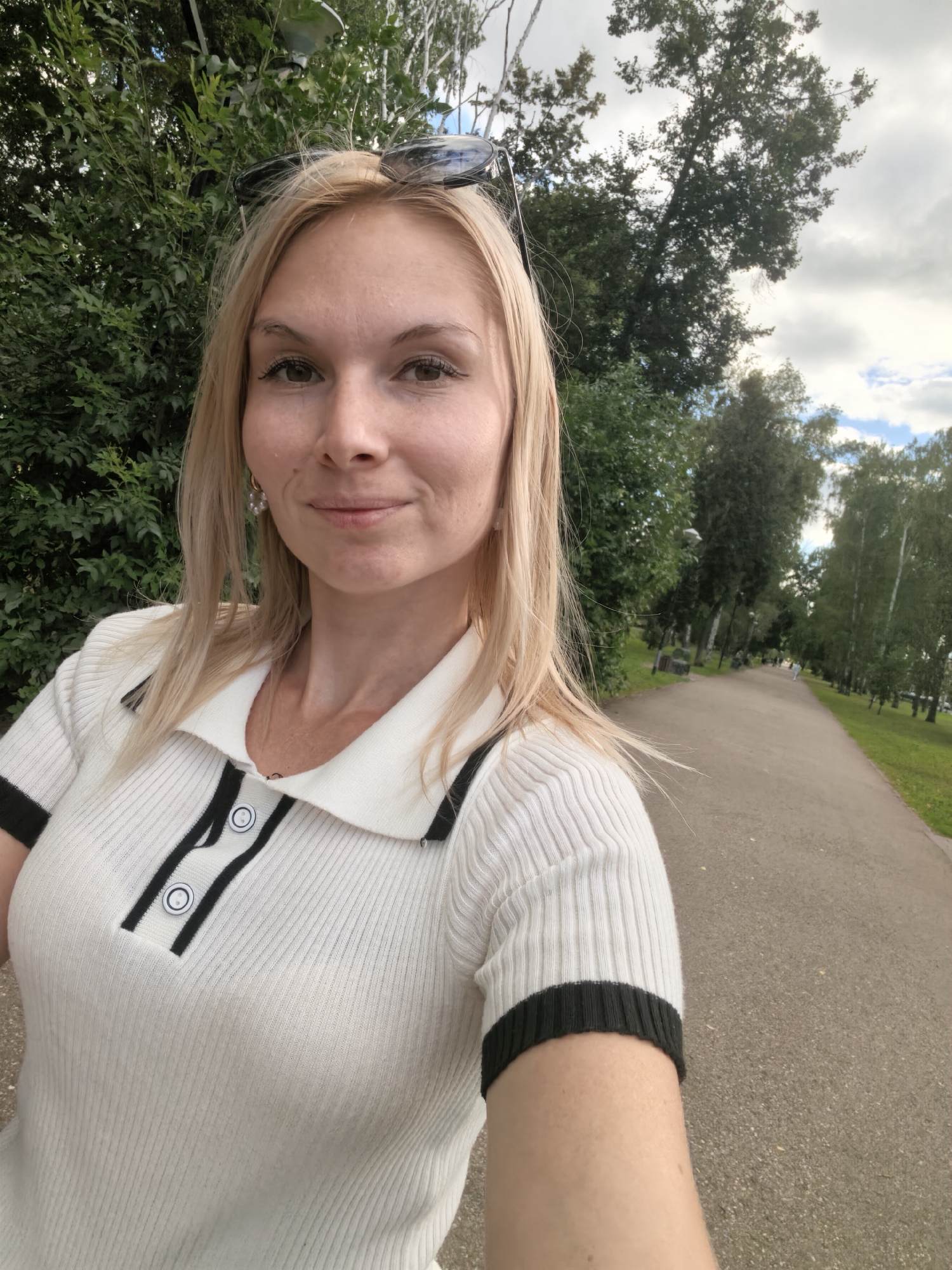 A blonde woman with sunglasses perched on her head takes a selfie outdoors. She is wearing a white ribbed polo shirt with black trim and two buttons. The woman is smiling, and her hair is slightly windswept. Behind her, a paved path extends into the distance, flanked by trees and green grass. The sky is partly cloudy. This image was taken on a pleasant day in Sterlitamak, Russia.