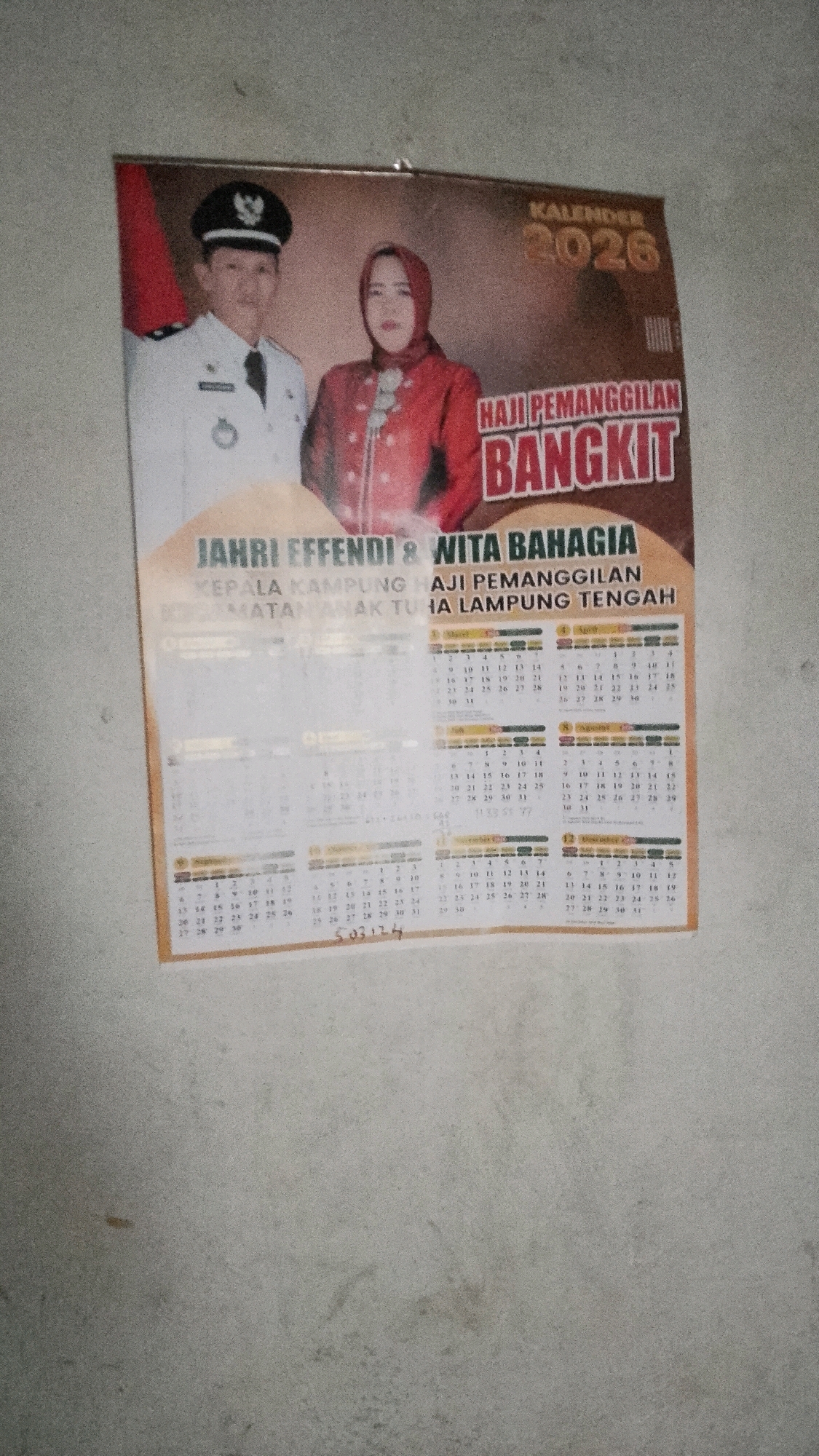 A wall calendar for the year 2026 is displayed on a light-colored, textured wall, likely in an indoor setting. The calendar features a portrait of a man and a woman. The man is wearing a white uniform with a black cap and a black tie, while the woman is wearing a red hijab and a red patterned top. Text above their heads reads "KALENDER 2026" and "HAJI PEMANGGILAN BANGKIT." Below the portraits, more text identifies them as "JAHRI EFFENDI & WITA BAHAGIA" and "KEPALA KAMPUNG HAJI PEMANGGILAN TAMAN ANAK TUHA LAMPUNG TENGAH." The calendar portion displays a grid of numbers representing days of the month for each month of 2026, arranged in columns and rows, with some text and small icons visible within some of the days. A handwritten number "503174" is present at the bottom of the calendar. The lighting is somewhat dim, casting a shadow on the lower portion of the calendar. The overall impression is of a political or community calendar.