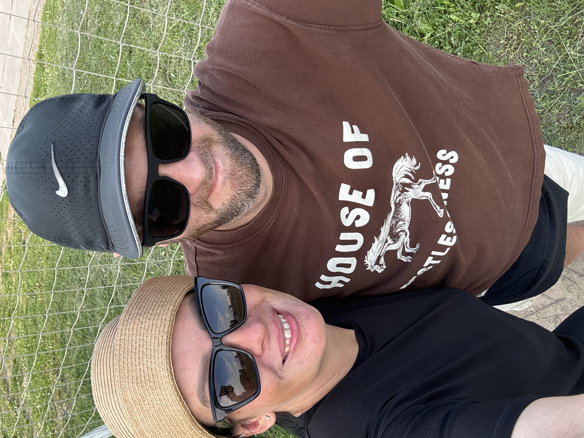 In a close-up, selfie-style photograph taken outdoors, a man and a woman are positioned side-by-side against a backdrop of a wire fence and green grass. The woman, on the left, wears a black t-shirt and a wide-brimmed straw hat, her face framed by large, dark sunglasses. She is smiling, with her head slightly tilted. A subtle silver earring is visible on her left earlobe, and a piercing is visible on her nose. The man, on the right, is wearing a brown t-shirt with the text "HOUSE OF" and a graphic of a horse printed on it. He is also wearing sunglasses and a black Nike baseball cap. Both individuals appear to be looking at the camera. The lighting suggests it is daytime. The exact location within Chaikovsky, Russia, cannot be determined from the visual information provided.