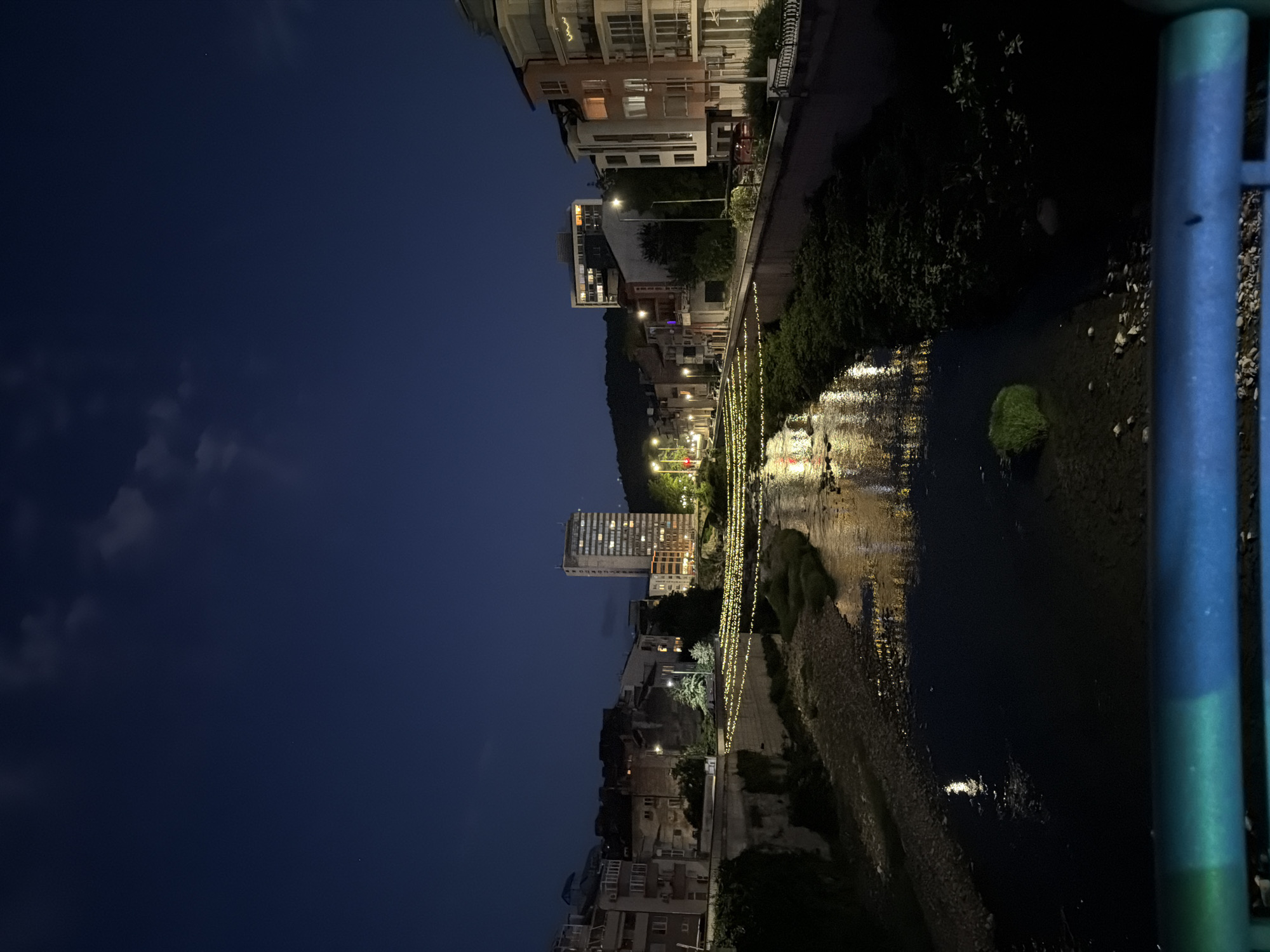 The image shows a night view of the Yantra River in Gabrovo, Bulgaria. String lights are draped across the river, reflecting on the water's surface. Buildings line the riverbanks, with some illuminated windows visible. A tall, modern building stands out among the other structures. The sky is dark blue with a few faint clouds and stars. The foreground features a blue railing.