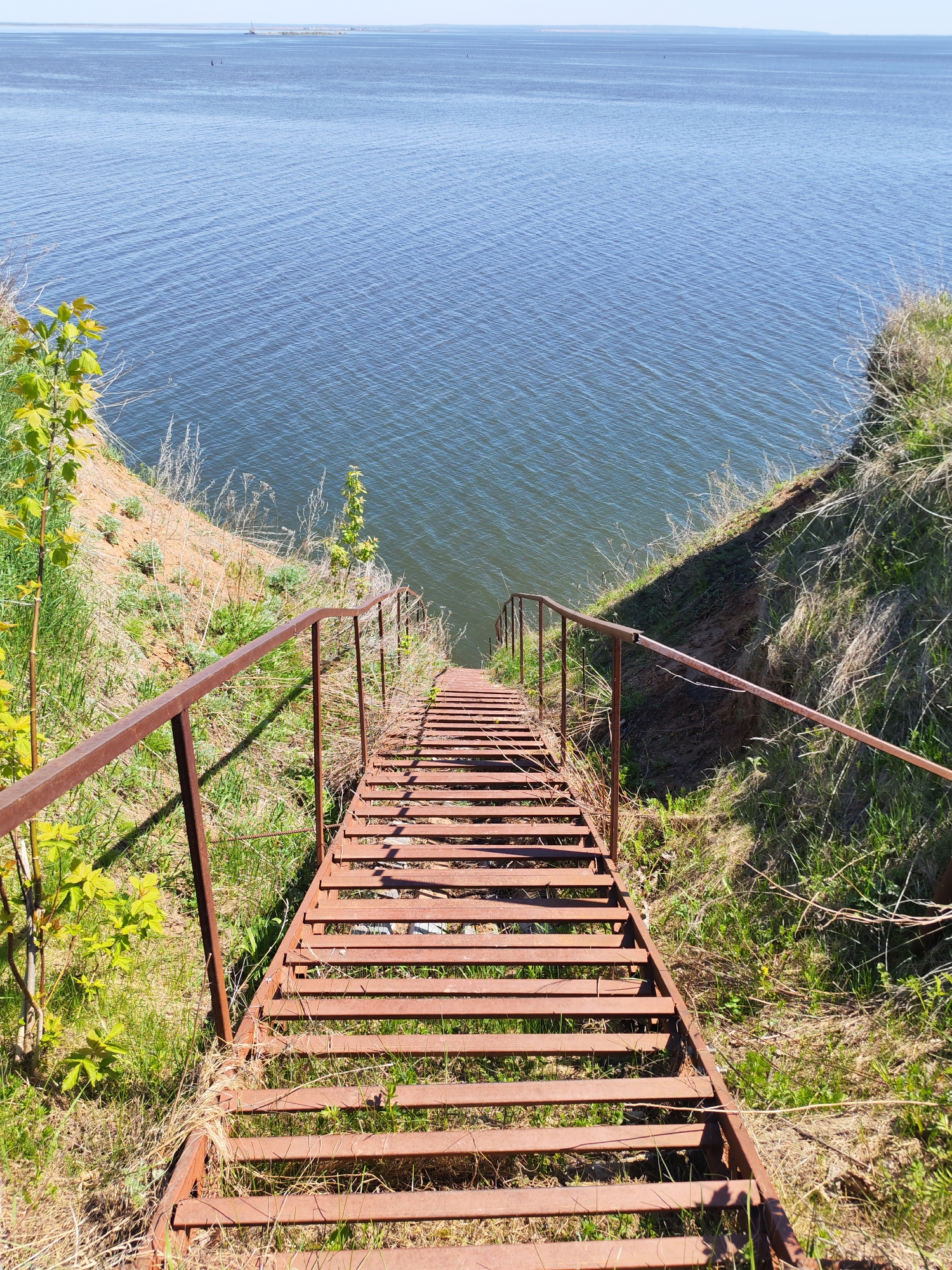 The image shows a rusty metal staircase leading down a grassy bank toward a large body of water. The water extends to the horizon, with a distant landmass visible on the far shore. The staircase has metal railings on both sides. The grass on the bank is mixed with some bare earth. Based on the location provided (Икское Устье, Russia) and the presence of a large body of water, it's likely the water body in question is a large river, lake or reservoir near this village. The image is taken from a high angle, looking down the staircase. The sky is clear and blue. There are no people visible in the scene.