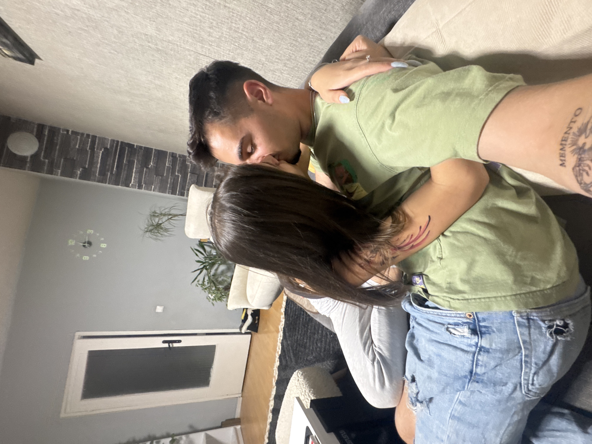 Two individuals, a man and a woman, are intimately embracing and kissing while seated or reclining on a sofa in an indoor living space, likely in Sofia, Bulgaria. The man, with short dark hair and some facial hair, wears a light olive green short-sleeved t-shirt. His right forearm features a tattoo clearly displaying the word "MEMENTO". The woman, with long dark hair, is dressed in a light-colored long-sleeved top and ripped light blue denim jeans. Her left arm, which is wrapped around the man's back, shows a tattoo of abstract red and purple lines near her shoulder. She is also wearing a ring with a clear stone on her left ring finger, and her fingernails are painted light blue.
The background reveals a home interior with a modern aesthetic, including a sofa, a wall adorned with dark textured panels, a plain light grey wall, and a white door with a dark panel. A glowing, circular wall clock is partially visible in the background, suggesting an indoor setting under artificial light, possibly in the evening or at night. Potted plants add a touch of greenery to the scene, which conveys a sense of affection and closeness between the two individuals.