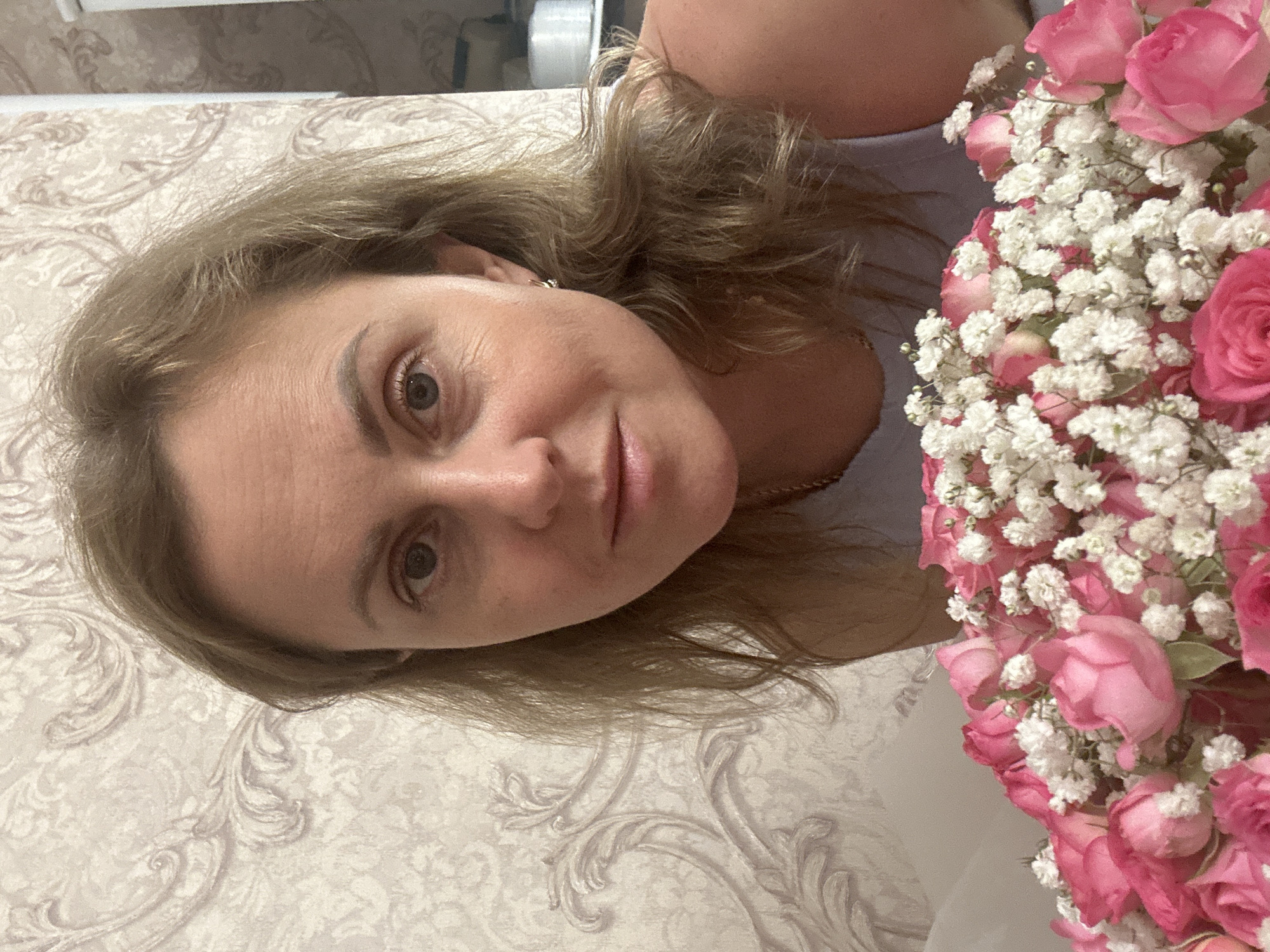 A woman with shoulder-length blonde hair is shown in a close-up portrait, holding a bouquet of pink roses and small white flowers. She is wearing a light blue top and a delicate gold necklace. Her expression is neutral, and she is looking towards the camera. The background is a light-colored wallpaper with a raised floral pattern. The image appears to be a selfie, taken indoors. There is no specific information about the location being in Verkhniy Yurt, Russia, in this image.