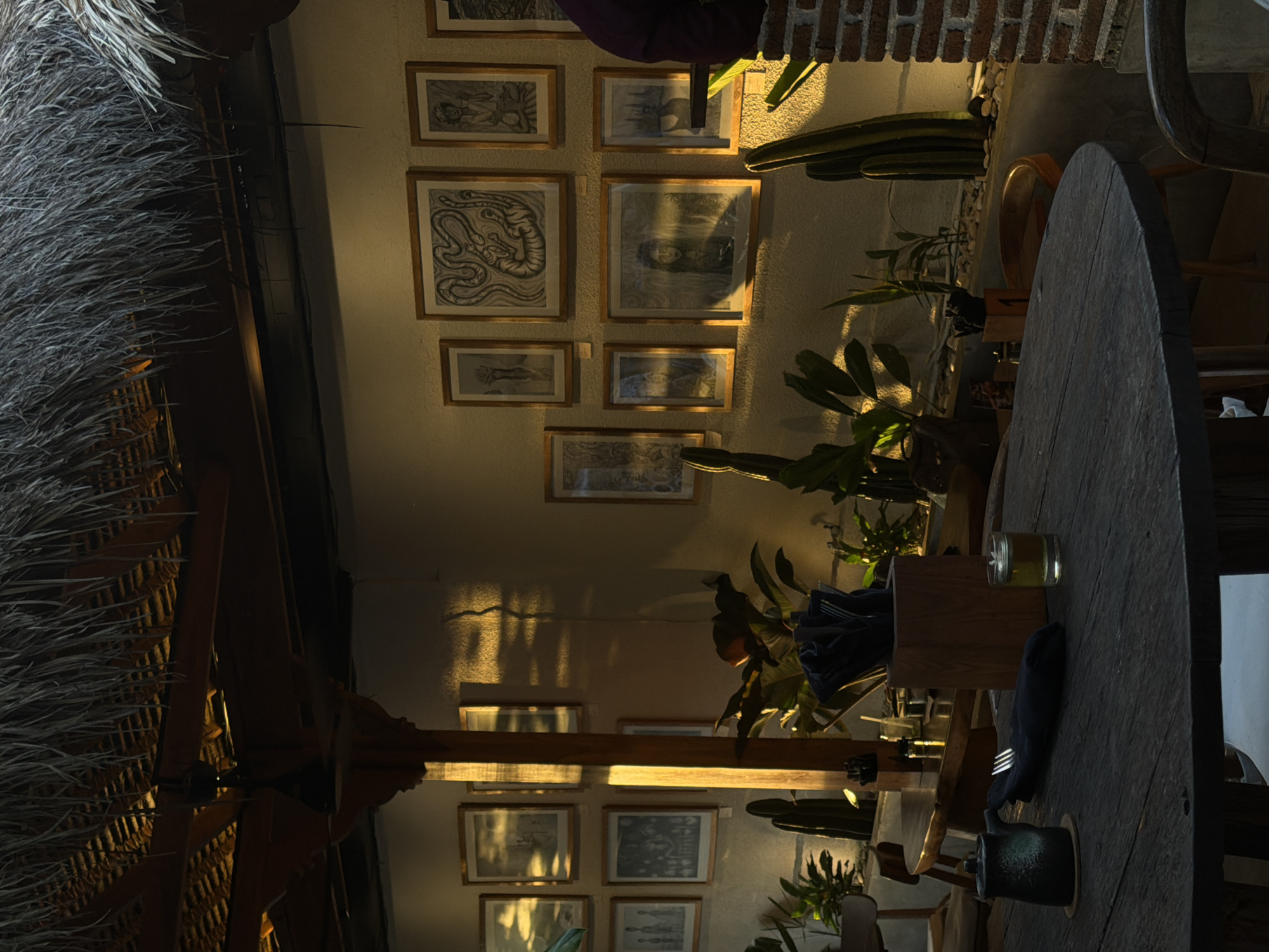 The image depicts an indoor dining space, seemingly within a restaurant or bar, in Kuta Selatan, Indonesia. The setting is under a thatched roof, lending a rustic and tropical ambiance. The walls are decorated with framed artwork, possibly prints or drawings, adding to the decor. Potted plants are scattered around the space, creating a lush and natural feel. A large, round wooden table is the centerpiece, set with napkins, silverware, and a small glass candle holder. The lighting appears warm and ambient, suggesting the scene is either during the late afternoon or evening. The focus seems to be on the ambiance and decorative elements of the dining space.