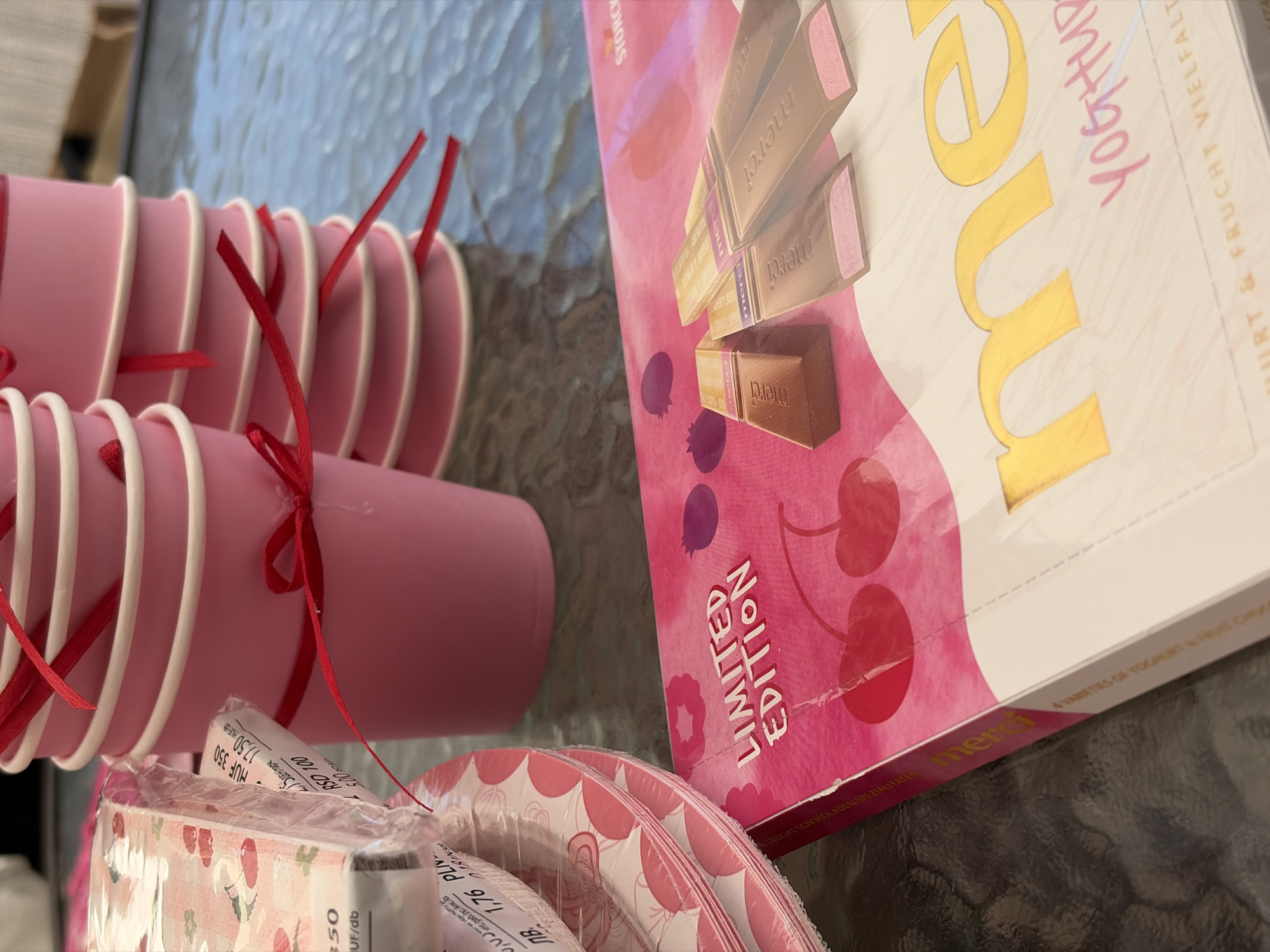 The image depicts a close-up of a table setting with pink paper cups and plates, a package of "merci" chocolates, and other party supplies. The pink paper cups are stacked and tied with red ribbon, creating a festive appearance. A stack of pink and white patterned paper plates is visible to the left, wrapped in plastic. Next to the plates, there is a package of "merci" chocolates, featuring a pink and white design with images of cherries and berries. The text "LIMITED EDITION" is prominently displayed on the chocolate packaging. Several individual "merci" chocolates are shown on top of the packaging, some with visible fruit fillings. The table surface appears to be made of glass, reflecting the surrounding objects. The overall scene suggests a celebration or party, with a focus on sweet treats and disposable tableware. The image does not provide any specific clues to its location within the city of Kolarovo, Bulgaria.