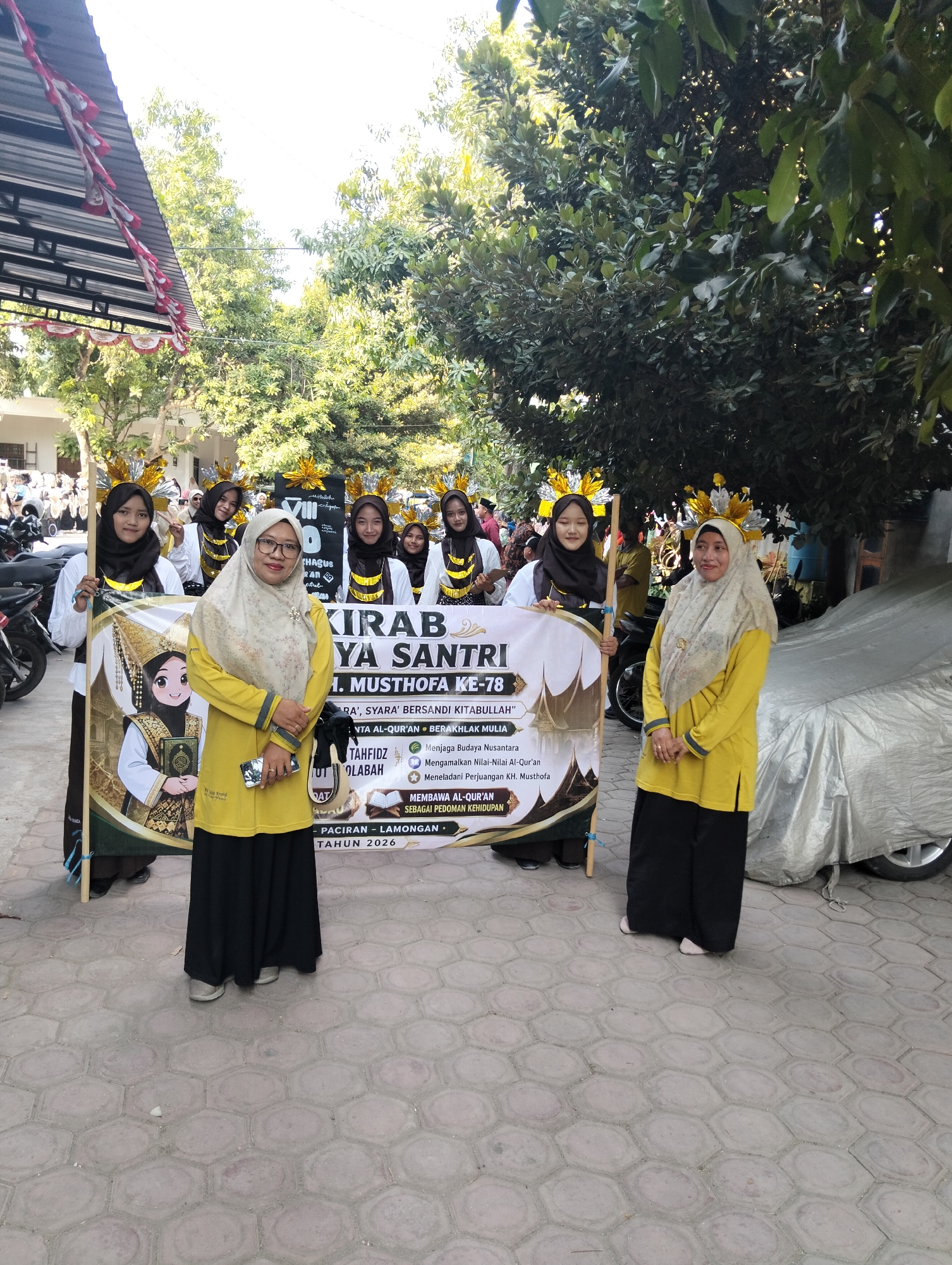 In Kranji, Indonesia, a group of young women dressed in black hijabs and white tops stand behind a banner. One woman, wearing a yellow hijab and yellow top, stands in front of the banner, holding a cell phone in her right hand. The banner reads "KIRAB YAKIN SANTRI K.H. MUSTHOFA KE-78" and has smaller text listing various religious and cultural organizations, along with the location "PACIRAN - LAMONGAN" and the year "TAHUN 2026". The women are adorned with gold headpieces. The background shows trees, a covered car, and buildings. The ground is paved with hexagonal tiles.