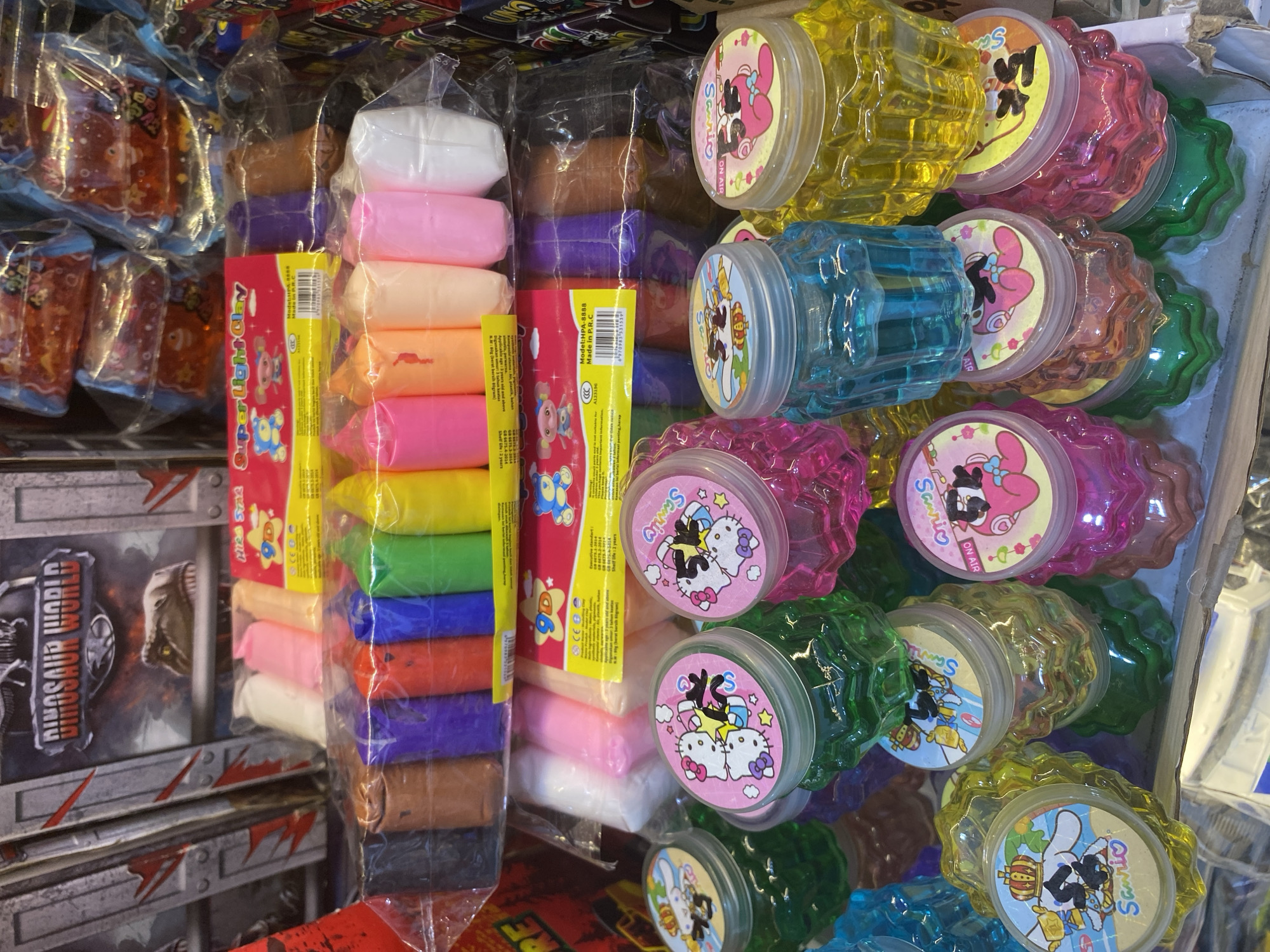 In a brightly lit retail environment, possibly a toy store or a market stall, an assortment of colorful children's toys is displayed. Dominating the foreground are several stacks of small, translucent plastic containers filled with brightly colored slime. These containers are topped with circular lids featuring various cartoon characters, including Hello Kitty and other Sanrio figures, often depicted in playful poses. The slime inside is visible in shades of green, pink, blue, and yellow.
Above the slime containers, packs of brightly colored modeling clay are visible. These packs are wrapped in plastic and contain multiple individually wrapped sticks of clay in various hues such as black, brown, purple, red, blue, green, and yellow, as well as pastel shades of pink, orange, and white. One pack prominently displays the text "Super Light Clay" and features cartoon characters.
In the background, a large cardboard box with "DINOSAUR WORLD" written across it is partially visible, along with imagery of a dinosaur's head. Other retail packaging and products are stacked behind the main displays, creating a sense of a crowded merchandise setting. The overall impression is one of a vibrant, toy-filled retail space designed to attract children. The scene does not provide specific clues to its exact location within Lamteumen Barat, Indonesia, but it is characteristic of a commercial area catering to toy sales.