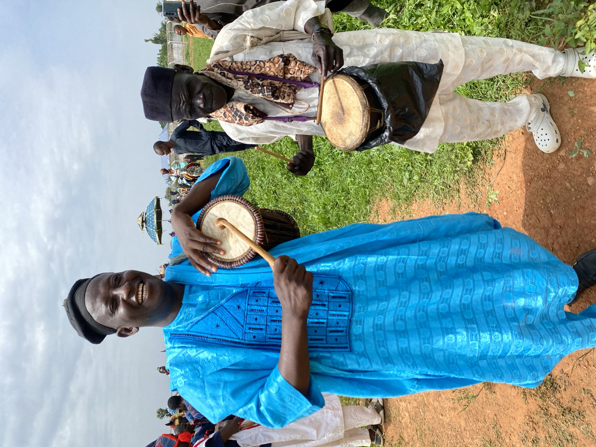 Two men, both wearing black hats, are playing drums. The man in the foreground is wearing a bright blue, traditional-style shirt, while the man behind him is wearing a white outfit with decorative embroidery. Both men are holding drumsticks and smiling. The scene appears to be outdoors, with green grass and a dirt path visible. The background includes other people, possibly spectators. The location is the city of Kafanchan A, Nigeria.
