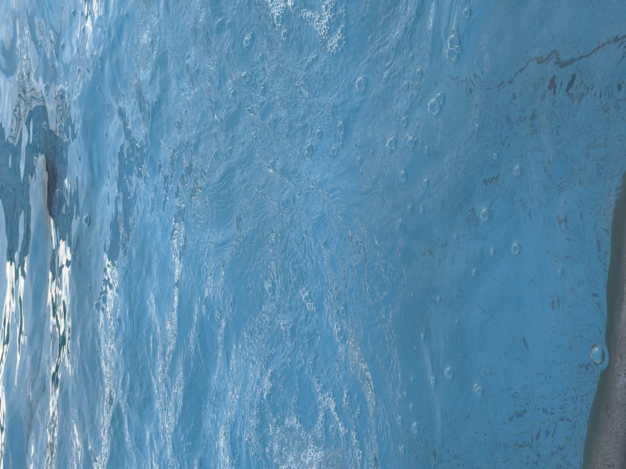 The image is a close-up, vertical shot of clear blue water with ripples and bubbles. The water appears to be in a swimming pool or a similar body of water. The lighting suggests it is daytime, with sunlight reflecting off the surface, creating bright highlights and casting subtle shadows. There are no people or identifiable objects in the frame, focusing solely on the texture and movement of the water. The bottom edge of the frame shows a slight hint of a gray surface, possibly the edge of the pool.