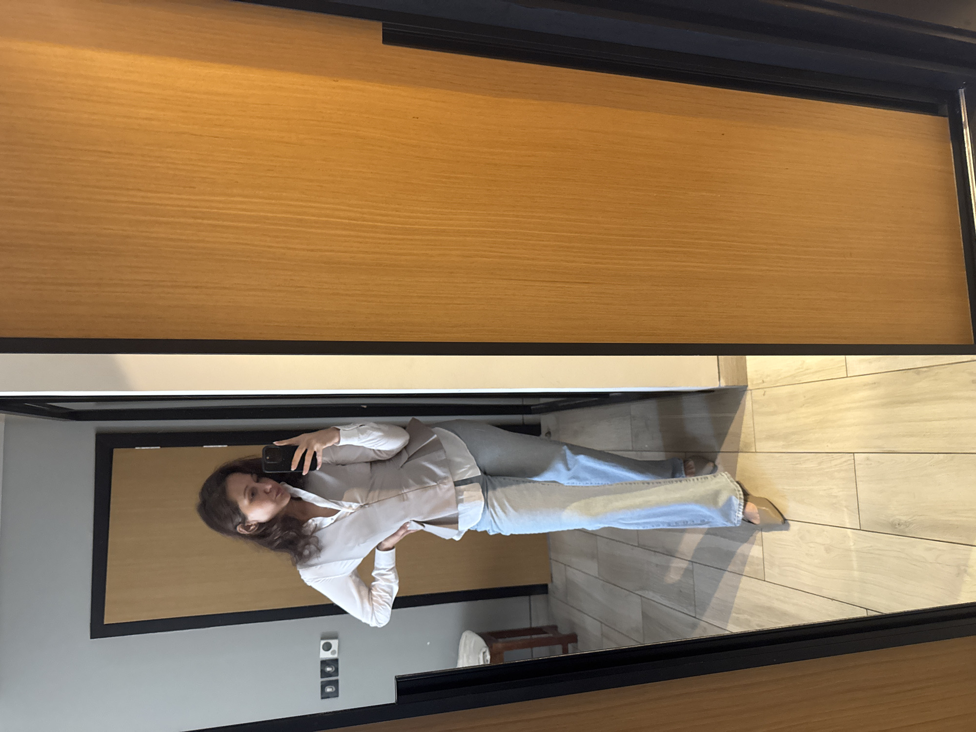 A woman with dark hair and wearing a white blouse and light blue jeans is taking a selfie in a mirror. The mirror is framed with a black border and reflects a portion of a room. The floor is light-colored wood and there is a small table, a wall with a wood door, and electrical outlets visible. It is not possible to confirm if the image was taken in Dubai, United Arab Emirates.