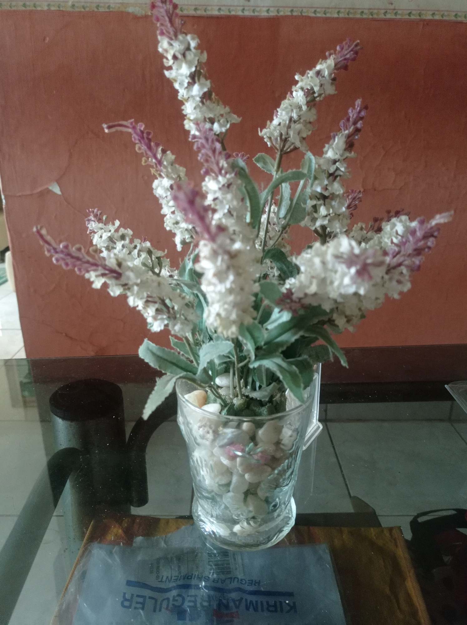 A close-up, eye-level shot features a bouquet of artificial white and lavender flowers in a clear glass vase filled with small white pebbles. The vase is positioned on a glass table. Behind the flowers and vase, a textured coral-colored wall serves as the backdrop. To the left of the vase, a dark, curved object, possibly a handle or part of a lamp, is visible on the table. Partially visible beneath the table, in the foreground, is a piece of plastic packaging with blue lettering that appears to be in Indonesian, possibly indicating a shipment label. The lighting is dim, casting some shadows. This scene is likely found in a domestic setting or a commercial establishment in Bandung City, Indonesia.