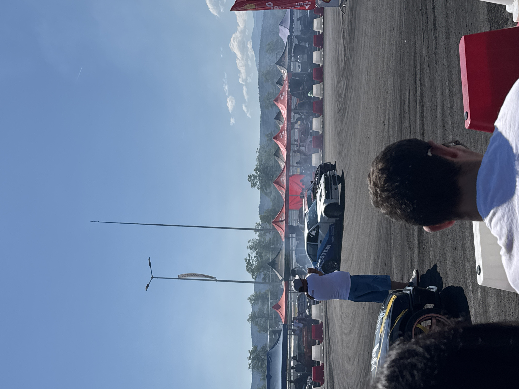 The image depicts a sunny day at an outdoor motorsport event, likely a drift competition, as indicated by the presence of smoke from tires and specially designed race cars. The scene is set on a dusty, dark asphalt track, with spectators visible in the foreground and mid-ground. Several tents, predominantly red and blue, are set up along the perimeter of the track, providing shade and likely housing teams or vendors.
In the mid-ground, two drift cars are actively engaged. One is a white sedan with blue accents and the text "Bilstein" visible on its side, emitting a significant amount of smoke from its tires as it maneuvers through a turn. Another car, partially visible in the lower left, is black with yellow accents and has a bright pink rim on its front wheel. A person in a white shirt and blue shorts stands near the white drift car, facing away from the camera and appearing to hold up a phone or camera, possibly recording the event.
The environment includes a clear blue sky with a few scattered clouds and distant, tree-covered hills or mountains. Tall light poles are also present on the track. The ground surface is rough and marked with tire tracks.
The specific location is stated to be the city of Dospey, Bulgaria. The event appears to be a public spectacle with organized infrastructure for participants and viewers.