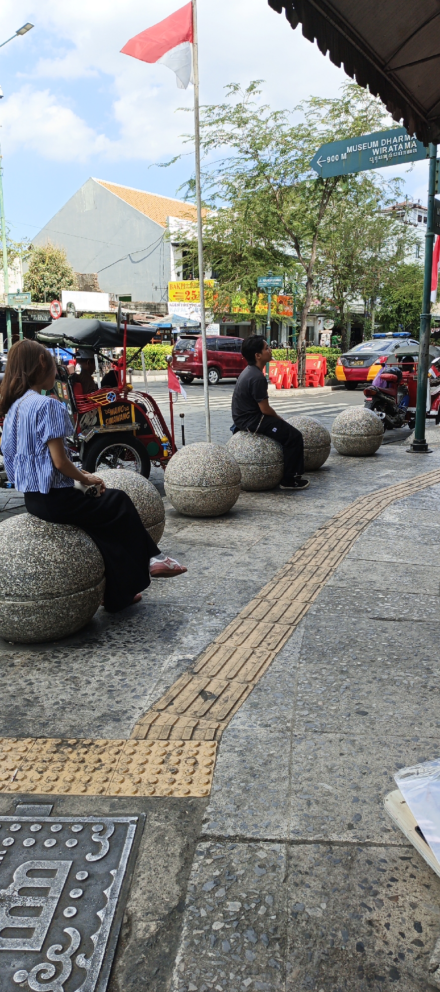 The image depicts a street scene in Yogyakarta, Indonesia, likely Malioboro Street, a well-known tourist area. In the foreground, two individuals are seated on large, round, grey stone benches. The person on the left, a woman with long brown hair, wears a blue and white striped blouse and black pants. The person on the right, a man wearing a black t-shirt and black pants, is looking up and to the right. Behind them, a colorful "becak" (a three-wheeled passenger vehicle) is visible, with the text "DEWANG 89 SINAWANG" on its side. Further in the background, a red minivan and other vehicles are on the road. The street is paved with a mosaic of grey stones, and a yellow tactile paving strip runs along the sidewalk, indicating it is a pedestrian area. A tall flagpole with the Indonesian flag (red and white) is prominently displayed, and a directional sign pointing to "MUSEUM DHARMA WIRATAMA" indicates a nearby attraction. Trees line the street, and buildings with orange-tiled roofs are visible in the distance. The sky is partly cloudy.