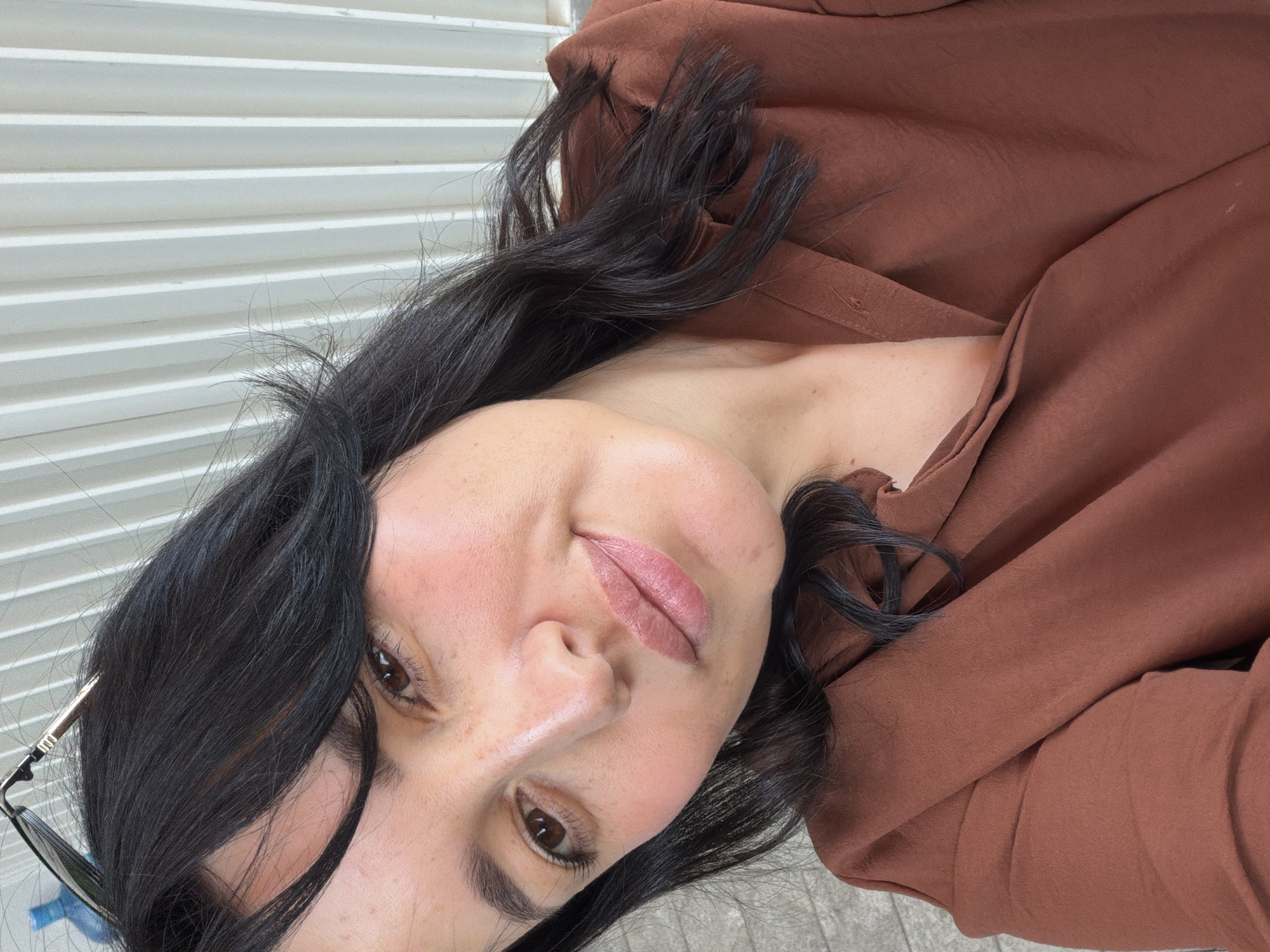 A selfie of a woman is taken outdoors, with a blurred background of a building. The woman has dark, shoulder-length hair and brown eyes. She is wearing a brown collared shirt and glasses perched on her head. The lighting suggests it is daytime. The background appears to be a neutral-colored corrugated metal wall.