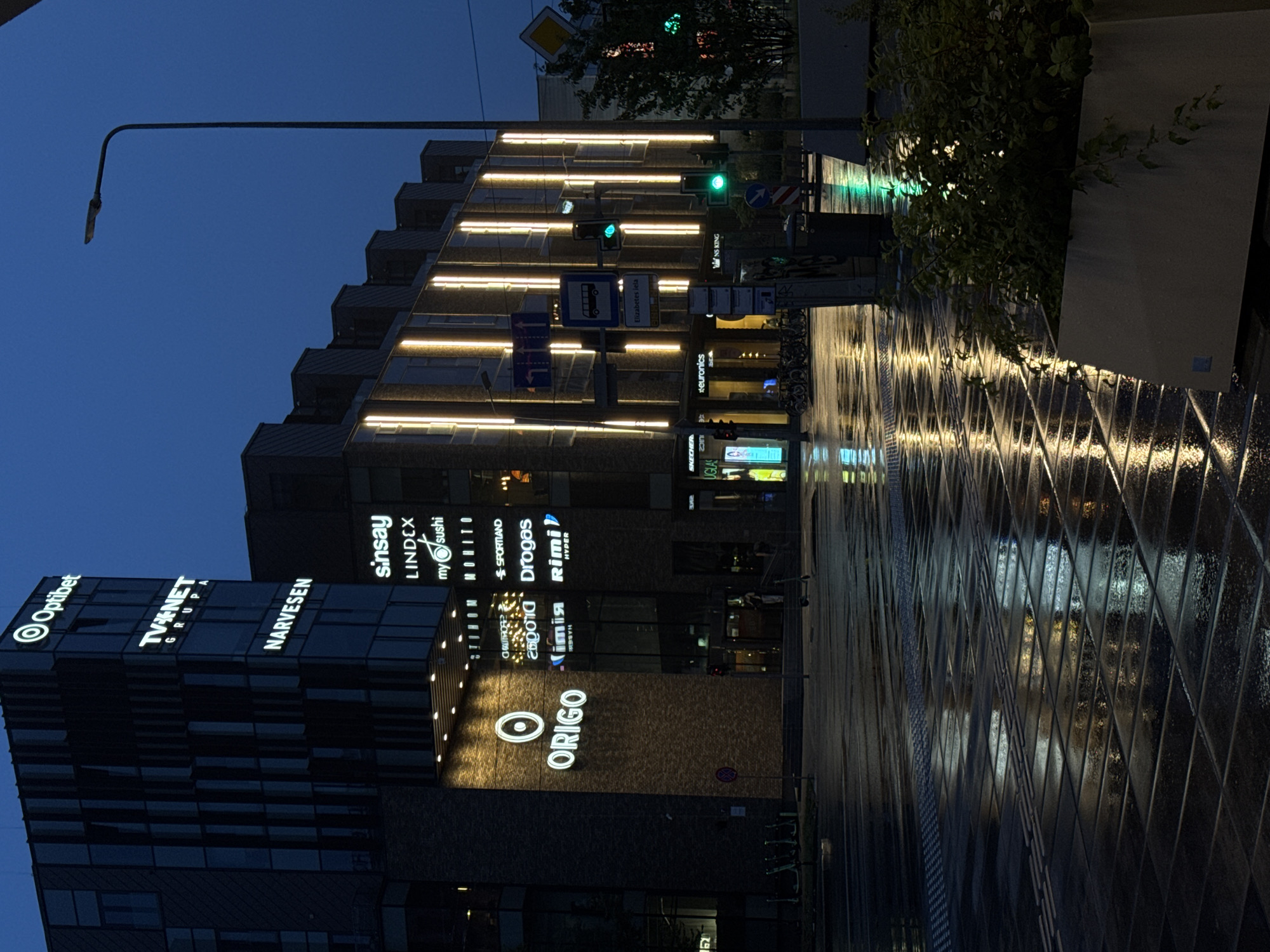 The image captures a street scene in Riga, Latvia, at dusk or night. The architecture of the buildings is modern, with dark facades and illuminated signage. Prominent signs include "Optibet," "TV NET GRUPA," and "NARVESEN" on one building, and "ORIGO" illuminated with a circular logo on another. Below "ORIGO," other signs like "SINOM," "MONITO," "SPORTLAND," "DROGAS," and "RIMI HYPER" are visible. The street is wet and reflective, suggesting recent rain, with lights from the buildings casting elongated reflections on the pavement. A lamppost stands to the right of the frame. Traffic signals are visible, with a green light illuminated. Several bicycles are parked on the left side of the street, and scooters are also present. A large planter with green foliage is situated in the foreground on the right. The overall atmosphere is urban and somewhat moody due to the low light conditions and reflections.