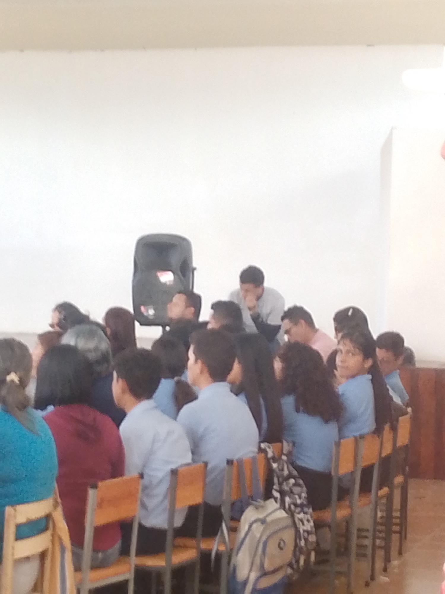 The image shows a group of people seated in what appears to be an indoor space in Juan Griego, Venezuela. Most of them are seated on wooden chairs with metal frames. Many of the people are wearing what appear to be school uniforms. A large speaker is located behind them. Some of the people appear to be listening to someone or something. A backpack is on the floor next to the chairs. The background is a plain white wall with a wooden panel on the right side.