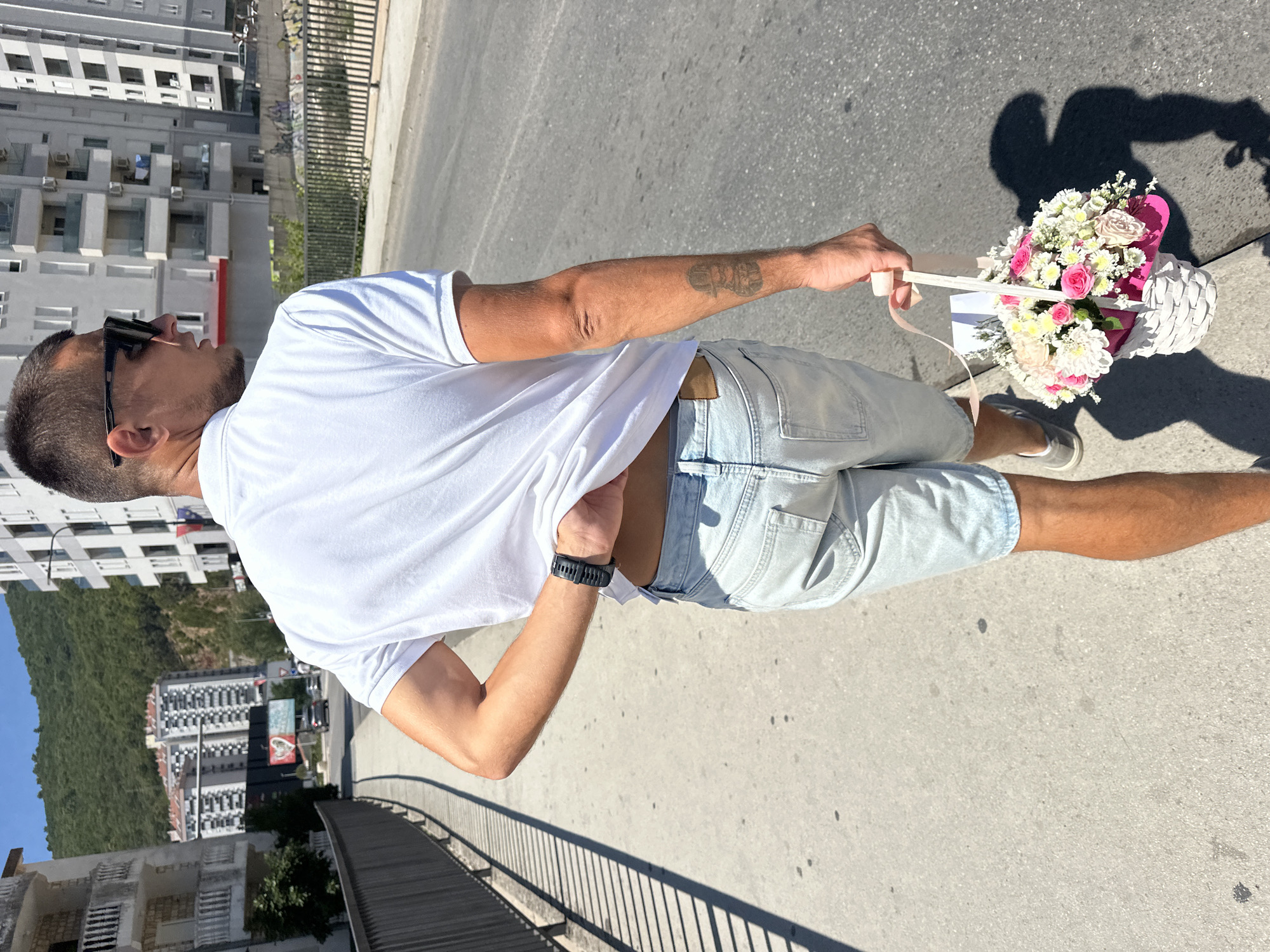 In Budva, Montenegro, a man walks down a sidewalk next to a road, carrying a white wicker basket filled with pink and white flowers adorned with a pink ribbon. He is wearing a white polo shirt, light blue denim shorts, and grey sneakers. The man's left arm is bent at the elbow, and his hand is tucked under the hem of his shirt, revealing a black wristwatch. He has dark hair, is wearing black sunglasses, and has a tattoo on his right forearm. The background features modern apartment buildings and a lush green hillside under a clear blue sky. The sun casts a shadow of the man and the flower basket on the concrete sidewalk.