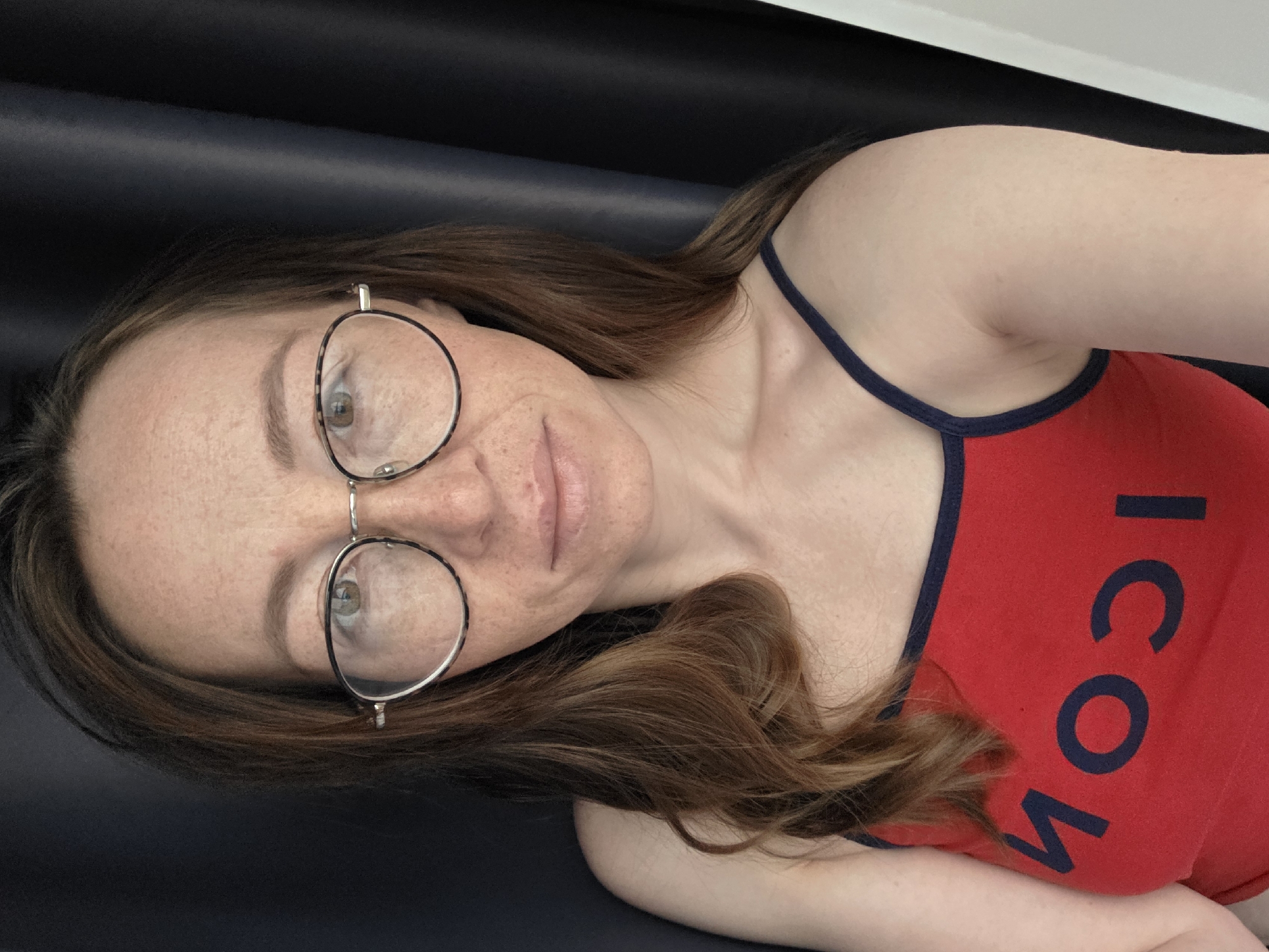 A selfie of a woman with freckles wearing glasses and a red tank top with blue trim and the word "ICON" printed in blue, lying on a dark blue surface. The background is a white wall. The location is not evident from the image.