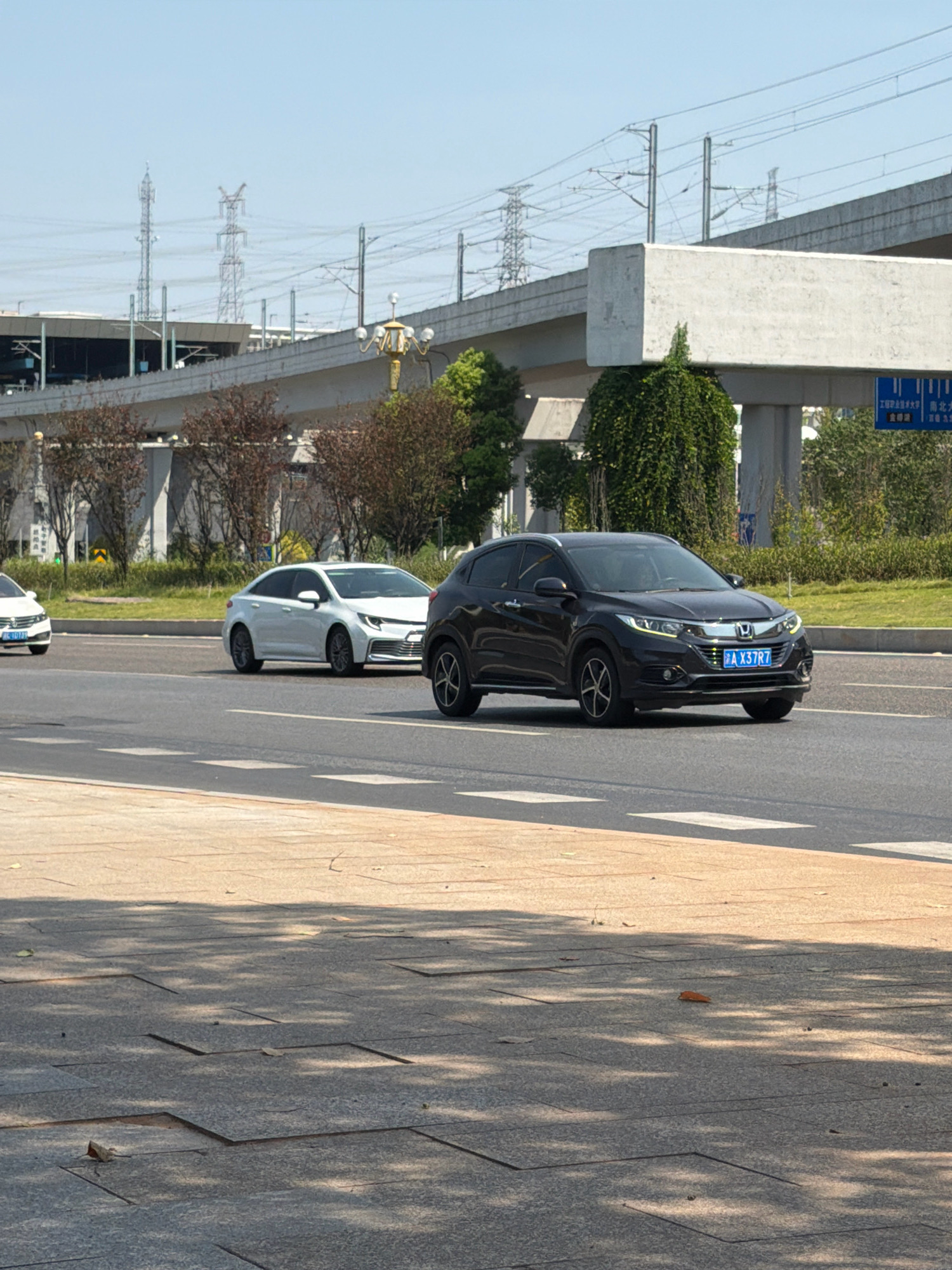 In Jiangjin, China, on a sunny day, cars are driving on a multi-lane road. In the foreground, a sidewalk is visible with large square tiles. In the middle ground, two white cars are driving on the road, one behind the other. The lead car appears to be a sedan, while the car behind it is an SUV. In the background, a black SUV is driving on the road. Beyond the road, there is a large elevated structure, likely a bridge or overpass, with electrical towers and wires. Green trees and shrubs line the sides of the road. A blue traffic sign is visible on the right side of the frame.