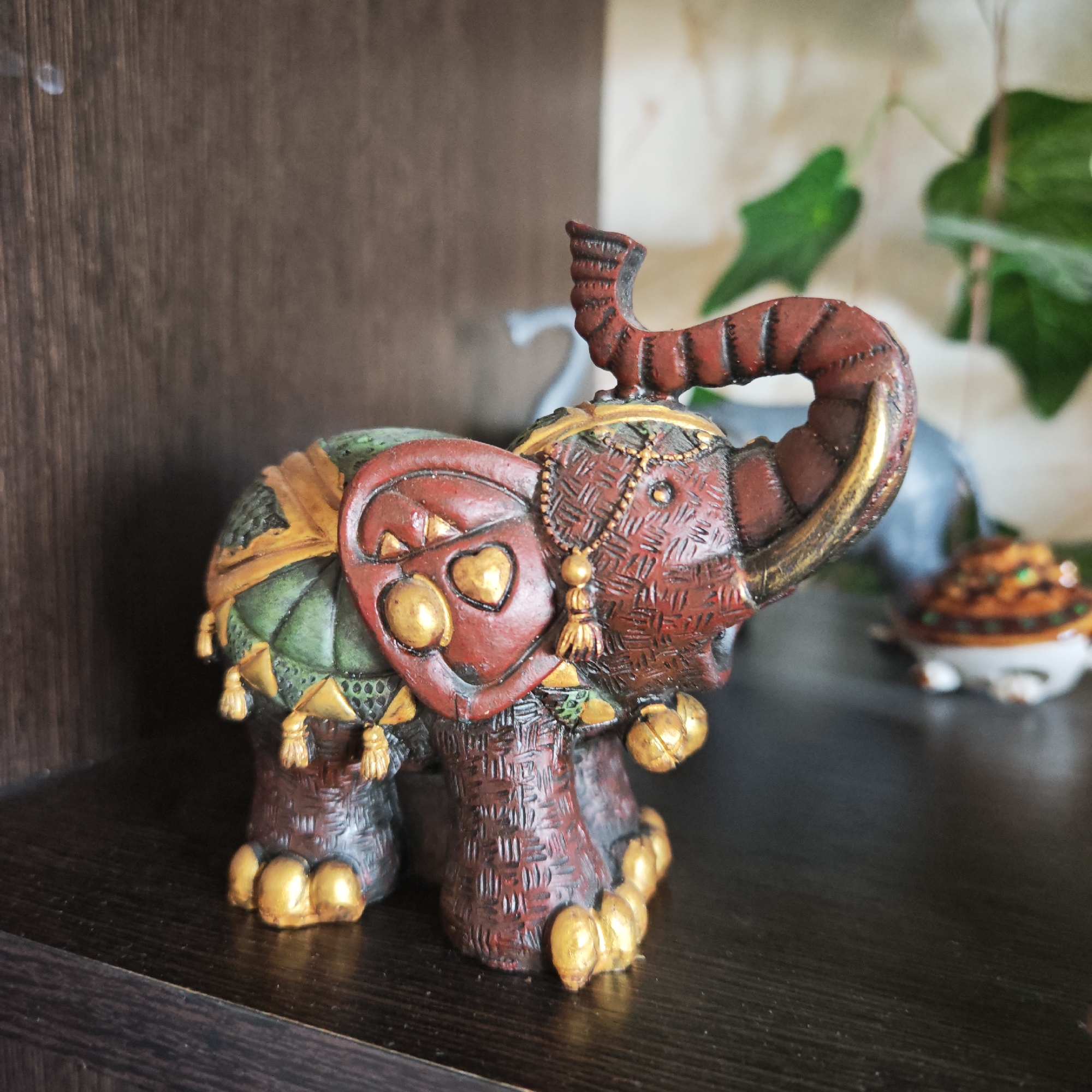 A brightly colored, intricately decorated elephant figurine is placed on a dark wooden shelf. The elephant is predominantly reddish-brown with gold accents on its feet, tusks, and ornamentation. Its trunk is raised. The elephant also has green and gold embellishments on its back and head. The shelf appears to be part of a dark wood cabinet or shelving unit. Behind the elephant, out of focus, are green leaves and what appears to be another figurine. The location is Rostov, Russia.