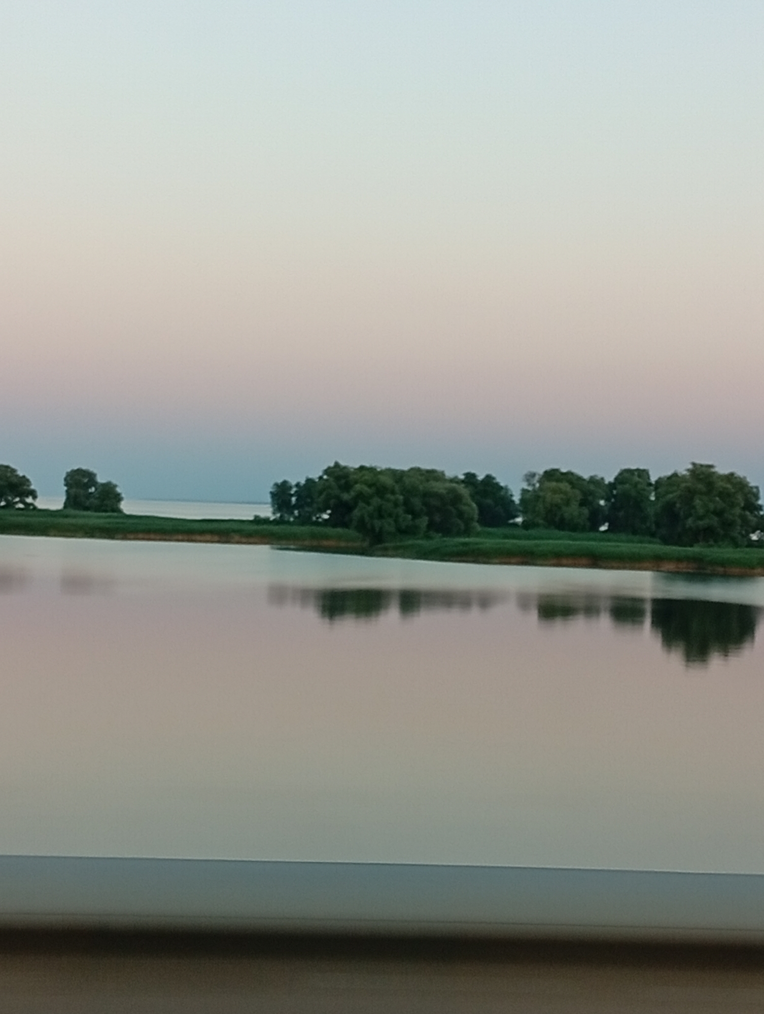 An outdoor, low-angle, eye-level shot captures a calm body of water reflecting a pale blue and pink sky at dusk. A strip of lush green land with scattered trees runs horizontally across the middle ground, bordering the water. The sky transitions from a light pink near the horizon to a pale blue at the top of the frame. The water is still, with clear reflections of the trees and the sky. In the foreground, a dark, horizontal structure, possibly a railing or barrier, is visible. The image does not contain any identifiable people or specific landmarks.