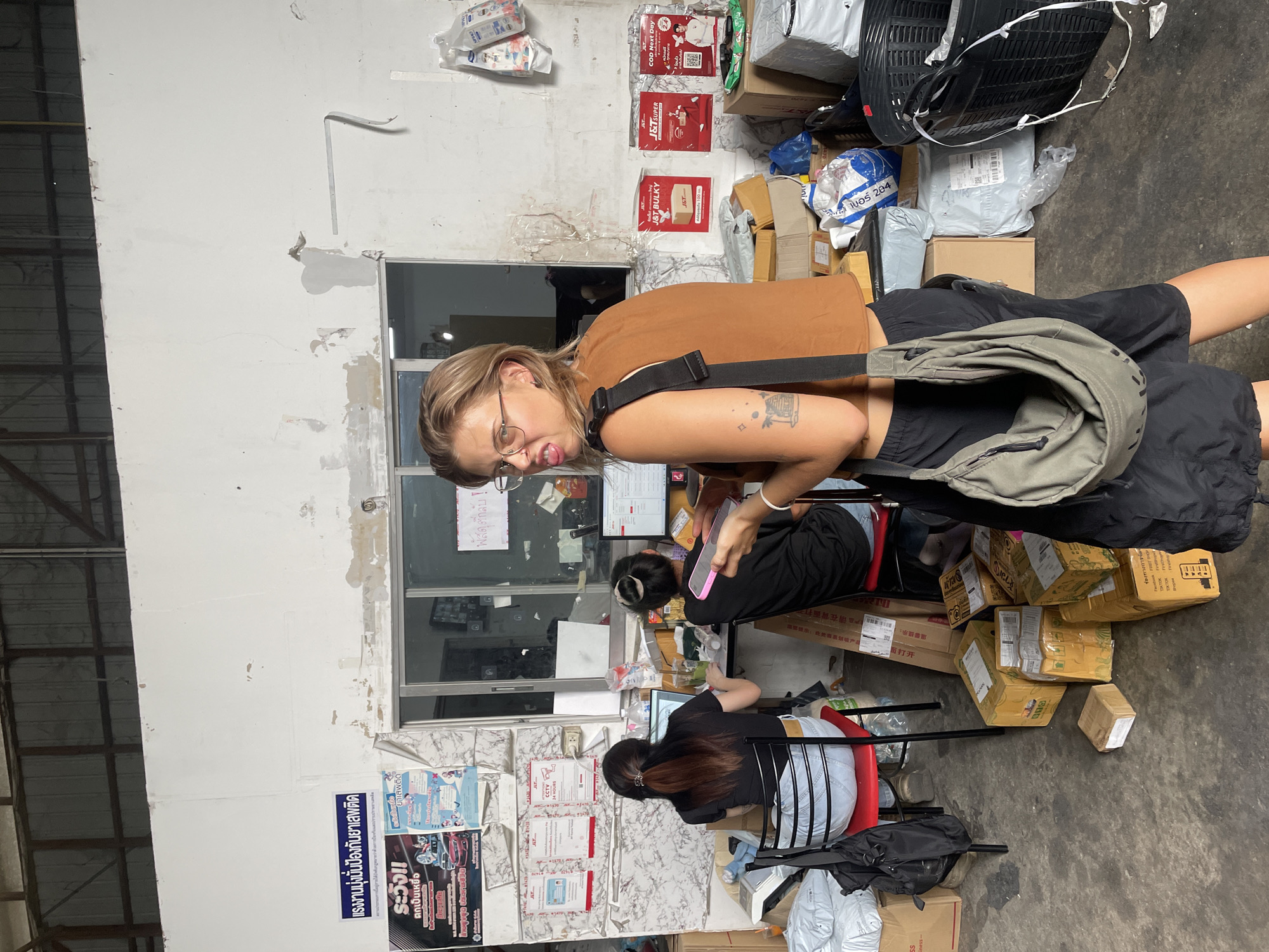 The image shows an indoor scene in what appears to be a shipping or mailing business located in Chiang Mai City Municipality, Thailand. There are three women present: one stands in the foreground wearing a brown top, black shorts, and a grey bag, while the other two are seated at a desk in front of a window. The seated women are facing computers and dealing with various packages. Numerous cardboard boxes and plastic parcels are scattered around them and stacked against the wall. The wall is cluttered with posters and notices in Thai script. A black fan, a large black basket, and more packaged items are also visible along the wall near the ceiling. The overall environment appears busy and somewhat cluttered.