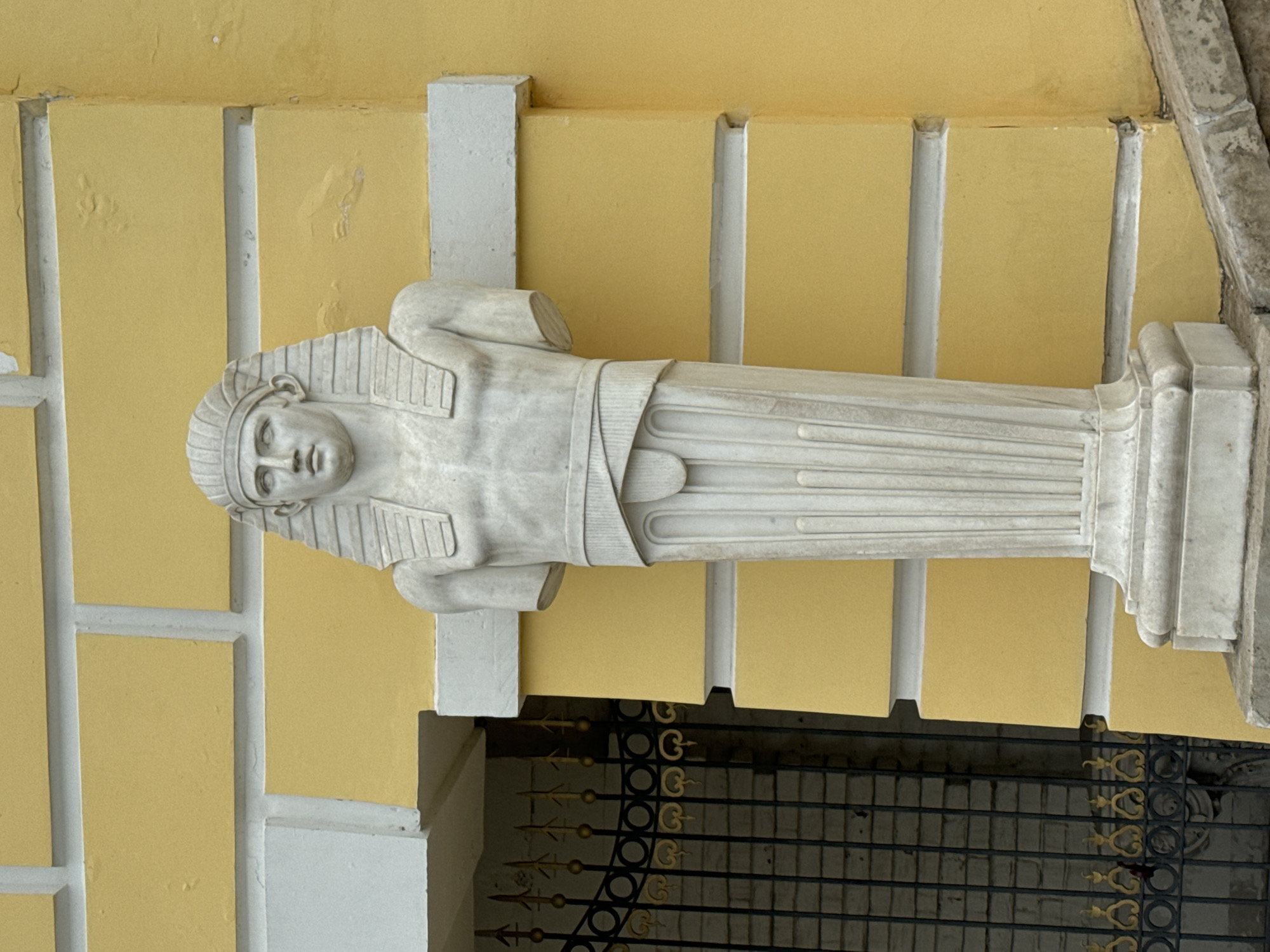 A horizontal, eye-level shot shows a white marble statue of a pharaoh mounted on the outside wall of a light-yellow building. The statue has a human face and head with a striped headdress, and its arms are crossed over its chest. The statue is wrapped in a white marble sheath that is striped vertically. The statue sits on a white base with vertical and horizontal striations. The building behind the statue is light yellow with vertical white columns. A section of black and gold fencing is visible beneath the statue. The building is located in the city of Arkhangel'skoe, Russia.