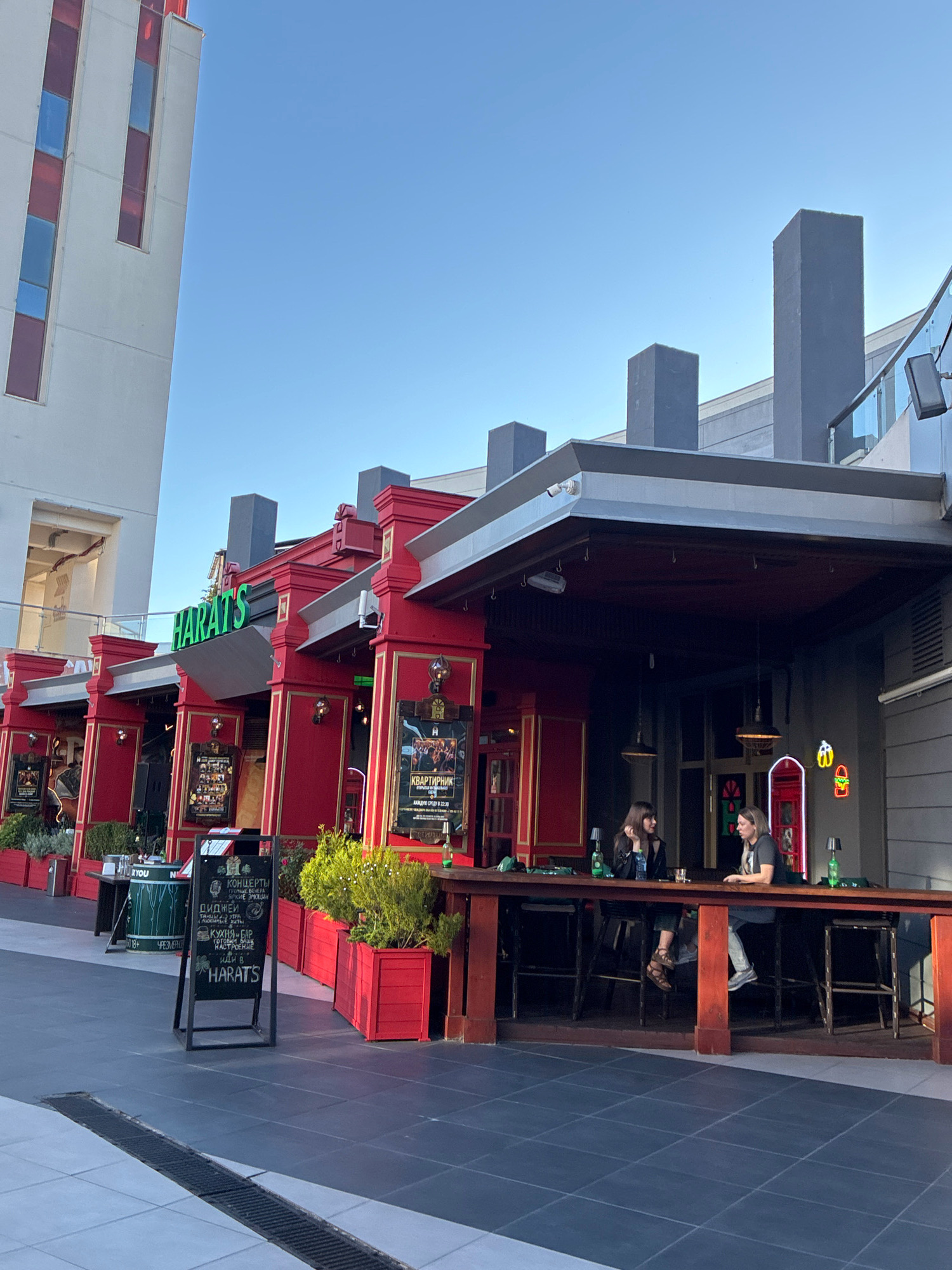 A bright, sunny day in Sochi, Russia, features the exterior of a red-and-gold building that appears to be a pub or restaurant named "HARAT'S." The building's facade is ornate, with prominent columns and decorative gold trim. Several people are visible, seated at a long wooden bar outside the establishment. Two women are seated at the bar, engaged in conversation. In the background, a modern, tall white building with red and blue rectangular window accents stands to the left, contrasting with the classic architecture of "HARAT'S." The ground in front of the pub is paved with dark gray square tiles. A black chalkboard sign displaying text in Cyrillic script stands near the entrance, along with potted green plants in red planters. Neon signs, including one depicting a hamburger, are visible on the building's exterior. The sky is clear and blue, indicating pleasant weather.