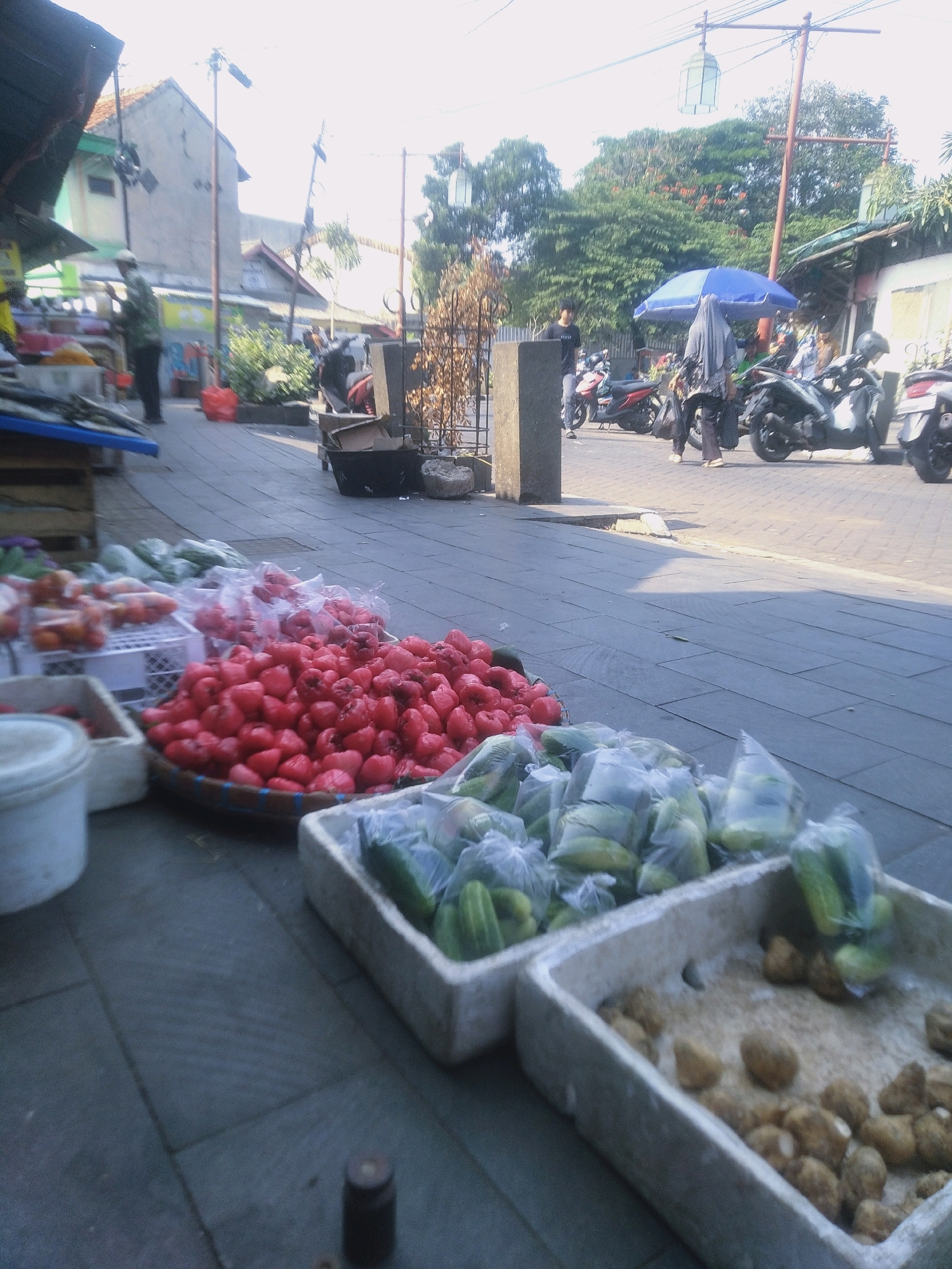 The image captures a bustling street scene in Cianjur, Indonesia, likely in a market area. In the foreground, various fresh produce is displayed for sale. A woven basket overflows with vibrant red fruits, possibly rose apples or wax apples, packed tightly together. To the right, a white rectangular container holds more red fruits, some of which appear to be wrapped in plastic. Adjacent to this, another white container is filled with green vegetables, predominantly cucumbers, also bagged in plastic. Further to the right, a similar container displays a collection of earthy-toned root vegetables, resembling potatoes or yams.
The backdrop reveals a street paved with interlocking gray bricks. Several motorcycles are parked along the side of the road, indicating a busy thoroughfare. A blue and white umbrella provides shade for a vendor or a customer, with a person wearing a headscarf standing nearby. In the distance, buildings with tiled roofs are visible, interspersed with lush green trees. A few individuals are seen walking or tending to their stalls. The lighting suggests it is daytime, with the sun casting shadows on the ground, hinting at a warm climate. The overall atmosphere is that of an active outdoor market where fresh produce is being sold.