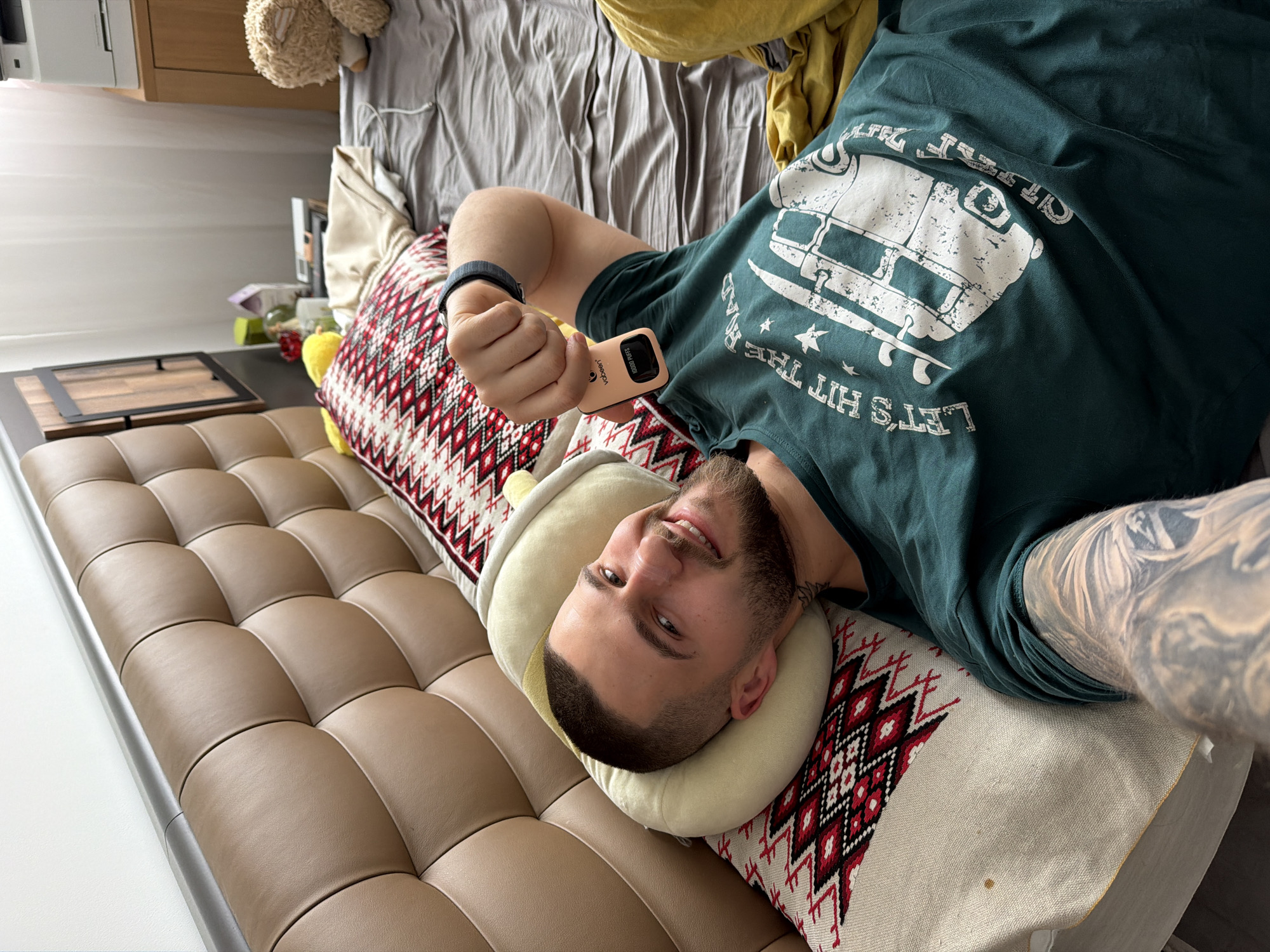 A bearded, light-skinned man lies on a brown, padded sofa in what appears to be an indoor setting. He is oriented with his head on the left and feet pointing to the right of the frame. He holds a peach-colored electronic device in his right hand, and a black smart watch is visible on his left wrist. He wears a dark green t-shirt with a white graphic of a van and the phrase "LET'S HIT THE ROAD" printed on it. His left arm features a full-sleeve tattoo. The man is supported by a beige neck pillow and a pillow covered with a geometric red and black pattern.
The background includes a bed with gray linens and a yellow object on top. A wooden cabinet is situated in the upper left, topped with a printer or similar device. Next to the sofa, there is a black framed object on a dark surface, along with other items, some of which appear to be food.
There is no indication of this picture being in Sofia, Bulgaria. The setting appears to be a private residence.