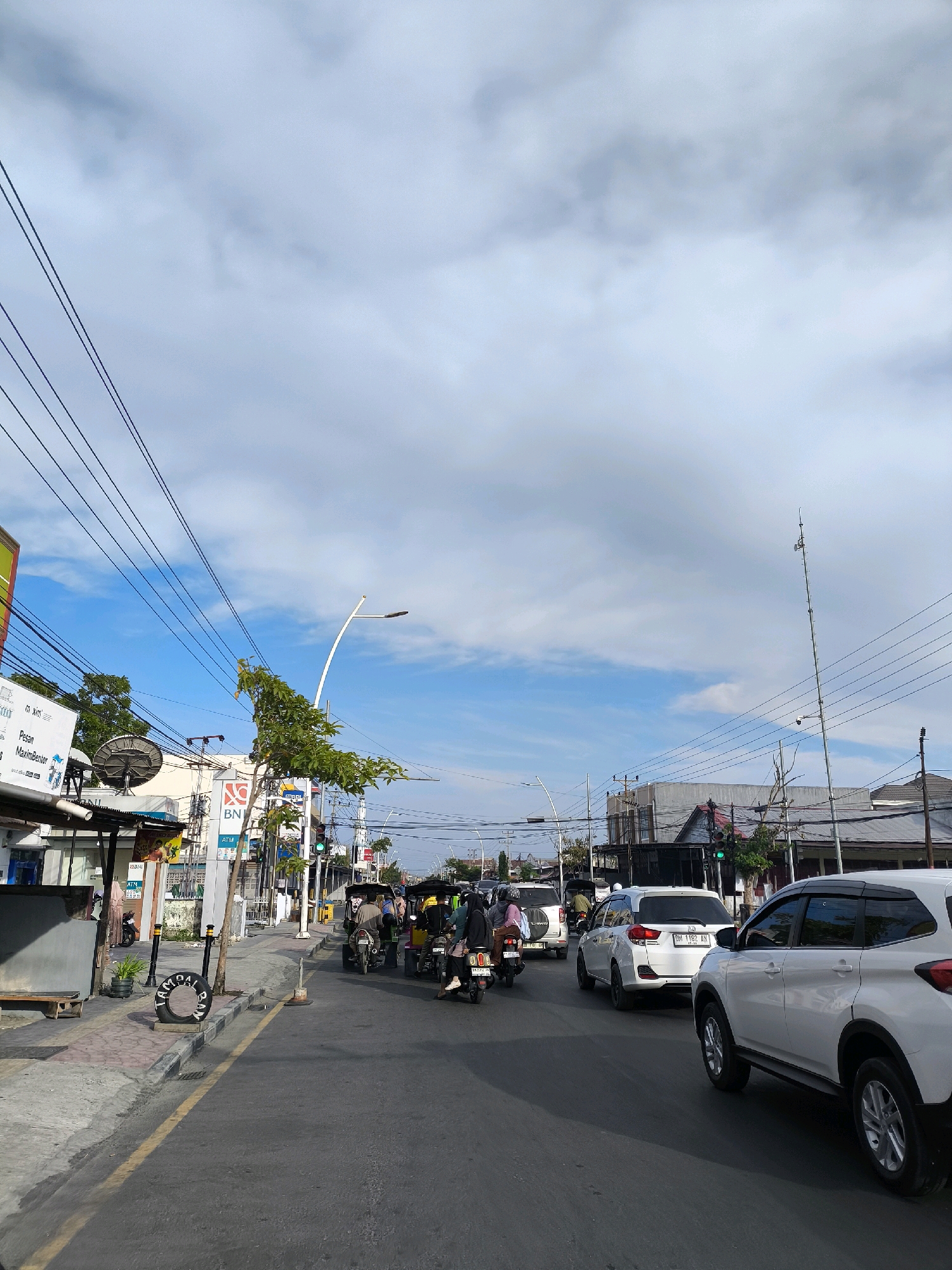 The image captures a street scene in Gorontalo, Indonesia. It shows a road with several vehicles, including cars and motorcycles, some of which appear to be tricycles. People are visible riding on these vehicles. The street is lined with buildings and commercial establishments, identified by signs that include "BNI" (a bank) and "ATM". Overhead, a complex network of electrical wires stretches across the sky, and several lampposts are present. Trees and foliage can be seen along the roadside and in front of some buildings. The sky is partly cloudy, with a mix of blue sky and white clouds. The overall impression is a busy urban environment on a bright day.