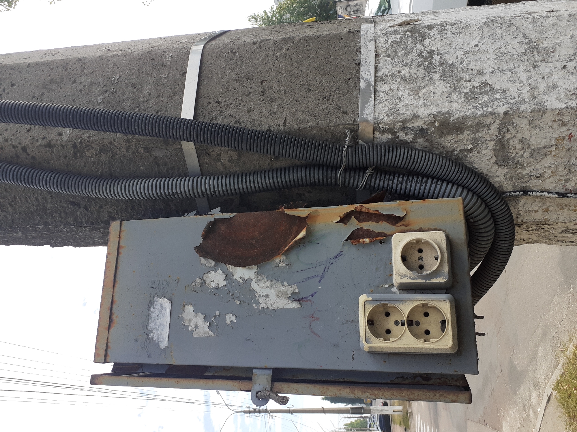 The image depicts an outdoor electrical junction box attached to a concrete pole on a street in Obukhiv, Ukraine. The gray metal box is weathered and shows signs of rust and peeling paint, with a large section of rust visible on the right side. Two electrical outlets are mounted on the front of the box; one is a double outlet, and the other is a single outlet. Both are light-colored and appear to be in functional condition despite the surrounding wear. Two black corrugated conduits run vertically along the side of the concrete pole, secured by metal bands. The pole itself is rough and textured, with patches of peeling paint or concrete. In the background, a street with cars and a pedestrian crossing is visible, suggesting an urban or suburban setting. The sky is overcast with some visible clouds. The scene is a candid shot, capturing a utilitarian piece of infrastructure in a somewhat dilapidated state.
