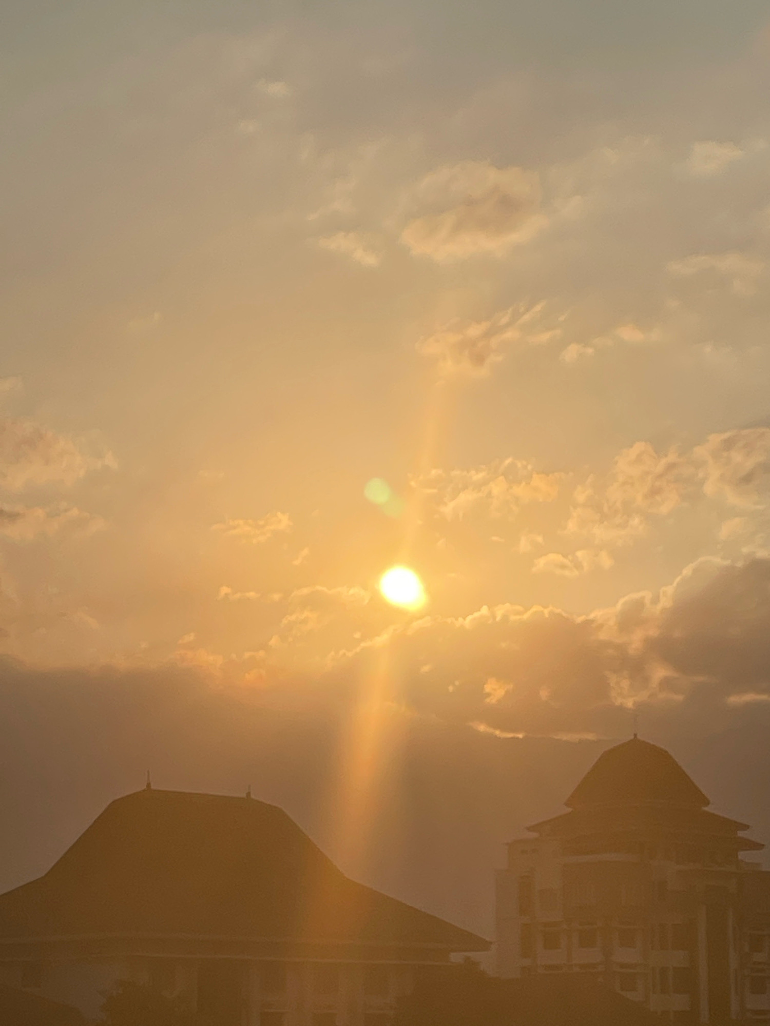 This image captures a sunset over a city landscape, likely in an urban or suburban setting. The sun is a bright, warm orb in the sky, casting a golden glow across the scene. It is partially obscured by scattered clouds, which are tinged with orange and yellow. The sun's rays are visible, creating a lens flare effect.
In the foreground, two distinct buildings are silhouetted against the vibrant sky. The building on the left has a distinctive, rounded, tiered roof, with what appear to be two small antenna-like structures at its peak. The building on the right is taller and more complex, with multiple stories, windows, and a prominent, conical or domed roof structure. Both structures appear to be architecturally significant. The lighting suggests it is late afternoon or early evening. The overall impression is one of tranquil beauty, with the warm light of the setting sun illuminating the urban structures.