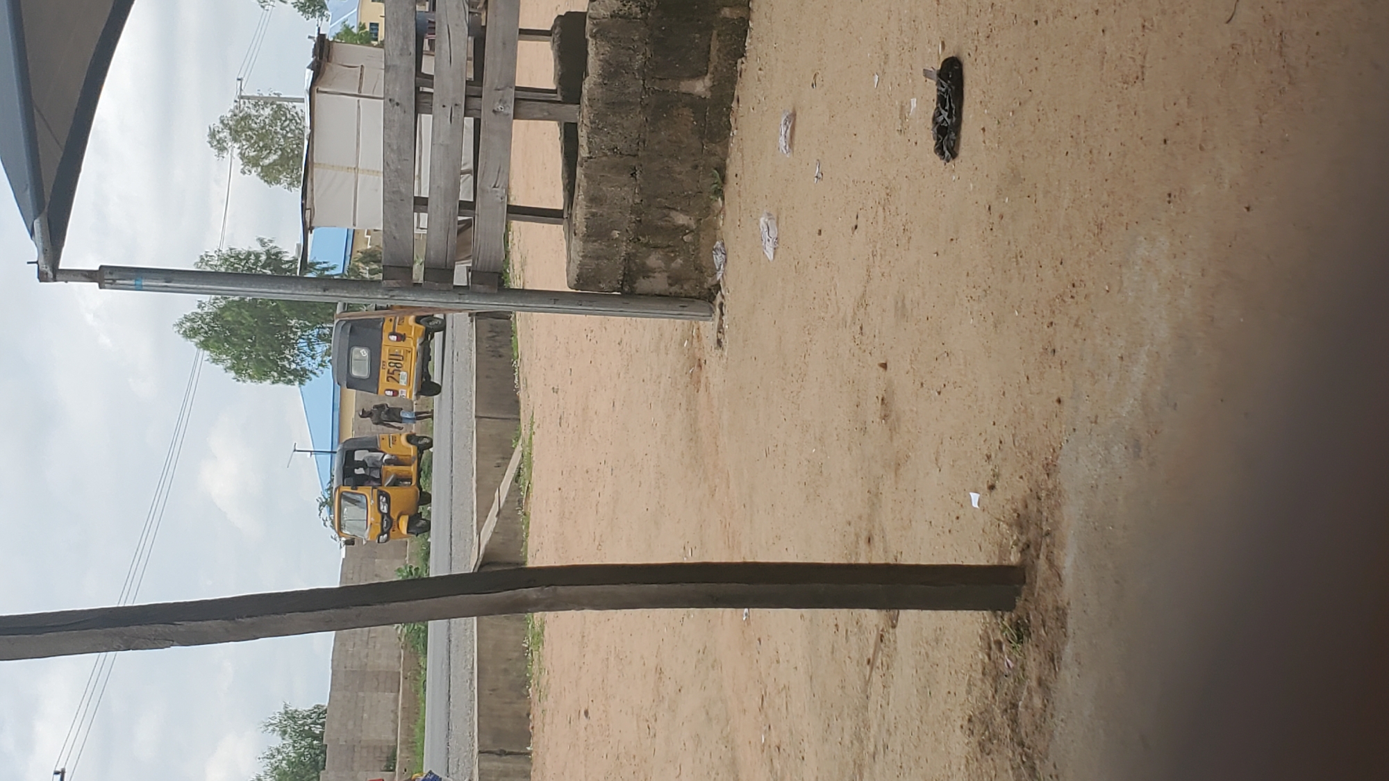 The image shows a dusty, unpaved area with a road in the background. Two yellow auto-rickshaws are on the road, one with two people inside and another parked. A person in dark clothing is standing beside the parked auto-rickshaw. In the foreground, a dark, vertical wooden post is prominent. A stone and wooden structure is visible on the right side of the image. The ground is covered with sand and scattered debris, including a black plastic bag on the right. The sky is overcast. The scene appears to be in a semi-urban or rural setting within the city of Jalingo, Nigeria.