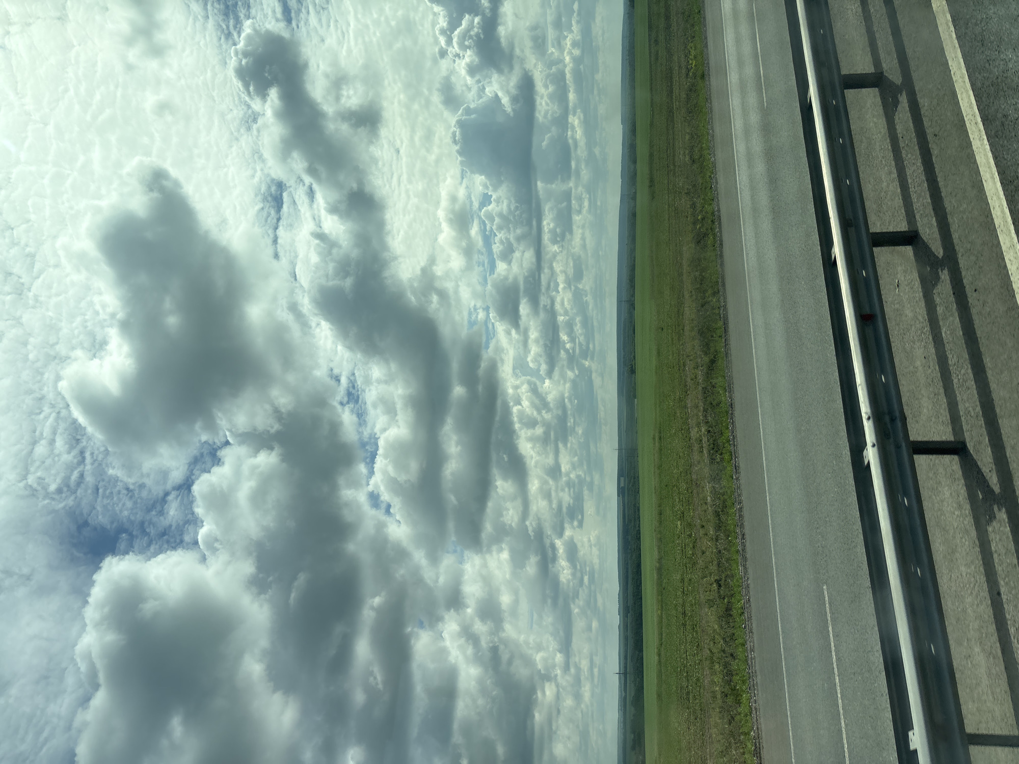 The image depicts a vast, open landscape under a dramatic, cloud-filled sky. In the foreground, a grey asphalt road runs horizontally across the frame, bordered by a metal guardrail with vertical supports casting shadows on the road. The road has a white dashed line marking its center. Beyond the road, a wide expanse of green fields stretches towards a distant, tree-lined horizon. The sky is filled with a complex arrangement of white and grey clouds, with patches of blue sky visible in between. The overall impression is one of a rural or semi-rural setting, possibly viewed from a moving vehicle. There are no people or specific city landmarks visible in the image that would indicate it is located in Menzelinsk, Russia.