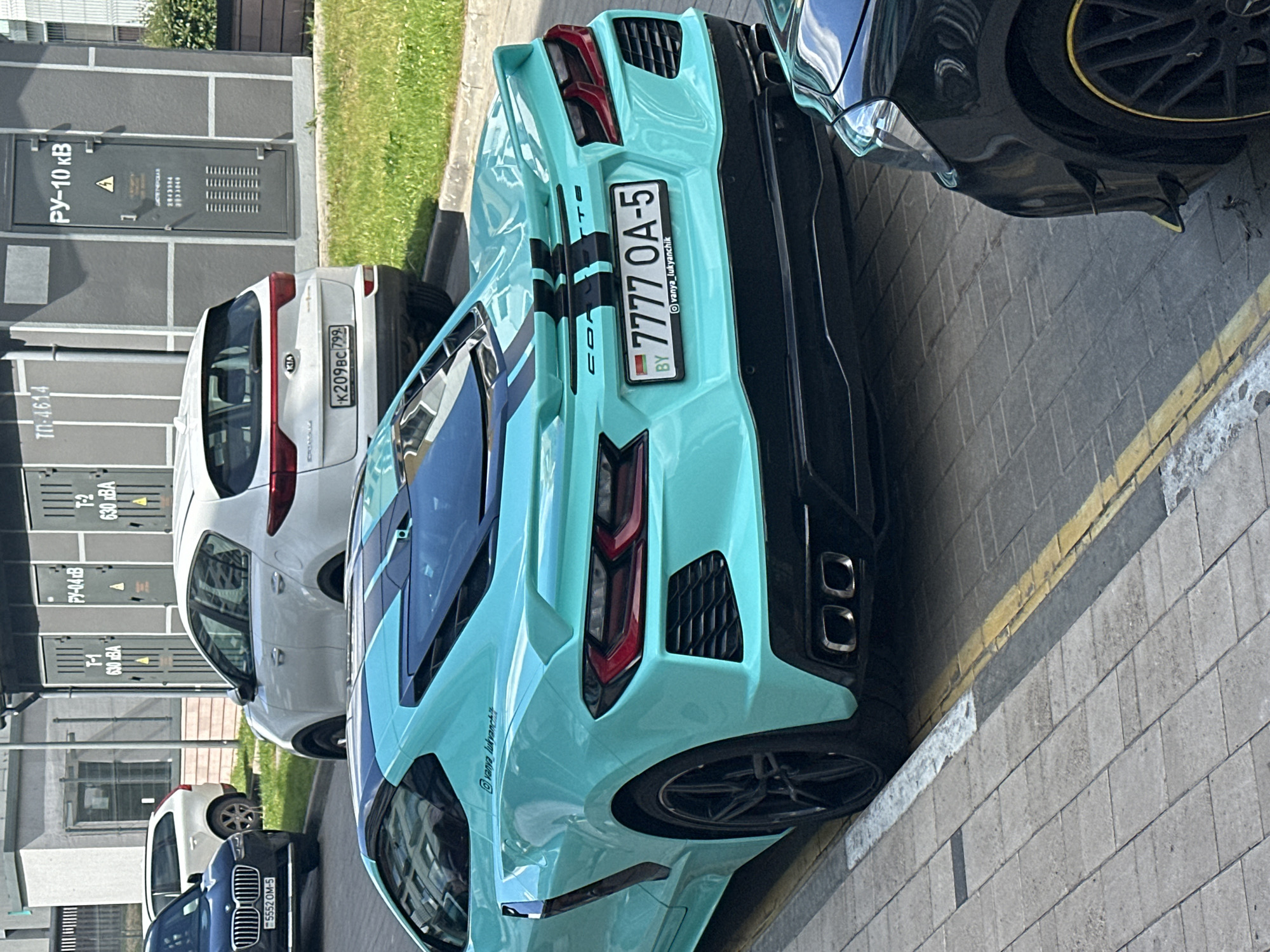 A daytime outdoor photograph captures a street scene in Бараўлянскі сельскі Савет, Belarus, featuring multiple parked vehicles and utility infrastructure.
In the foreground, a teal Chevrolet Corvette C8 is parked on a paved surface adjacent to a sidewalk, facing away from the viewer. The Corvette features black racing stripes running down its trunk and roof, black multi-spoke wheels, and a rear diffuser with two prominent square exhaust tips. Its Belarusian license plate reads "BY 7777 OA-5" and includes the social media handle "@vanya_lukyanchik" on the frame. A smaller "@vanya_lukyanchik" inscription is also visible on the rear quarter panel. A portion of a black vehicle, possibly a Mercedes-Benz with a yellow accent on its wheel rim and a canard on its front bumper, is visible in the bottom right corner, parked in front of the Corvette.
Behind the Corvette, a white Kia Sportage (fourth generation) is parked on the street. Its Russian license plate displays "K209BC799". Further behind the Kia, a black BMW X5 (F15 generation) is parked, partially visible on the left. Its Belarusian license plate reads "BY 5552 OM-5".
The background features a row of grey, rectangular utility cabinets or buildings. From left to right, visible labels include "ТП-4614", "Т-1 630 кВА", "РУ-10 кВ" (with a hazard symbol), and "Т-2 630 кВА". These labels denote transformer substations and switchgear, indicating electrical infrastructure. The ground consists of a grey tiled sidewalk with a yellow painted curb line and patches of green grass between the vehicles and the utility structures. The scene is illuminated by bright daylight, casting subtle shadows. No individuals are visible.