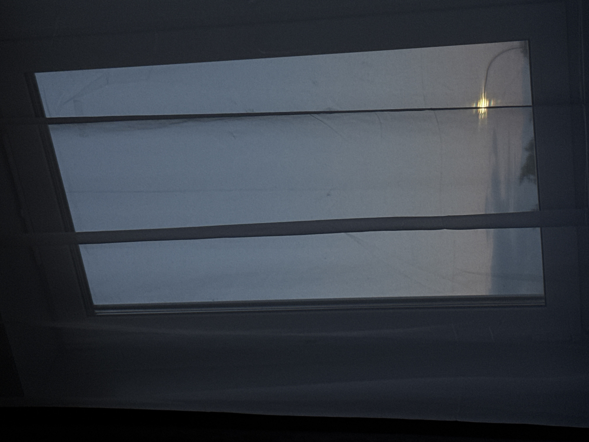 A close-up, slightly hazy shot captures a window with sheer white curtains partially drawn, revealing a muted, overcast sky. The sky appears to be twilight, with a hint of a streetlamp's reflection visible in the lower right pane of the window. The overall ambiance is subdued, with the light suggesting either dawn or dusk. The scene lacks any specific identifying landmarks that would pinpoint a location within Kaluga, Russia. There are no people, animals, or distinct objects other than the window, curtains, and the subtle reflection.