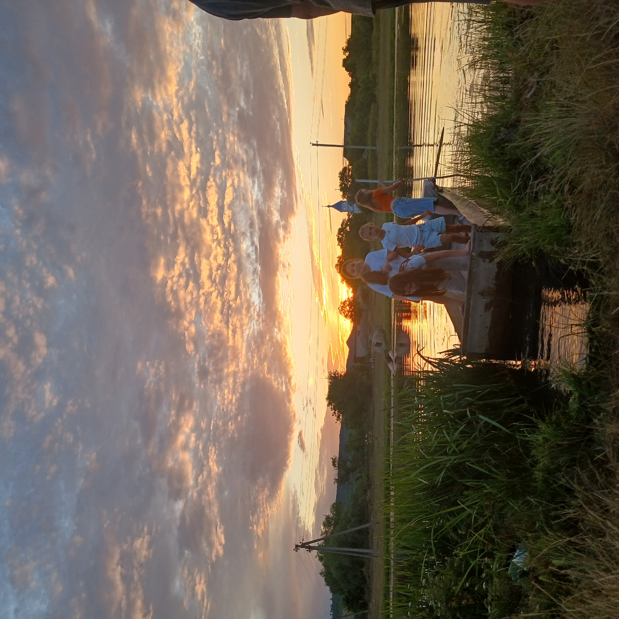 In Căzănești, Moldova, a boat carrying five children navigates a tranquil body of water during a sunset. The sky above is a dramatic canvas of orange and pink clouds, illuminated by the setting sun. The children, appearing to be of various ages, are positioned within the boat, some standing and others leaning over the edge. The boat itself is rustic, possibly made of wood, and rests low in the water. Surrounding the waterway is lush green vegetation, including tall grasses and reeds that line the banks. In the background, a treeline with rolling hills forms the horizon, and a distant church steeple with a blue dome is visible. A utility pole with wires stands in the middle ground, suggesting the proximity of human settlement. The overall scene depicts a peaceful, natural setting at dusk with the activity of children enjoying the water.