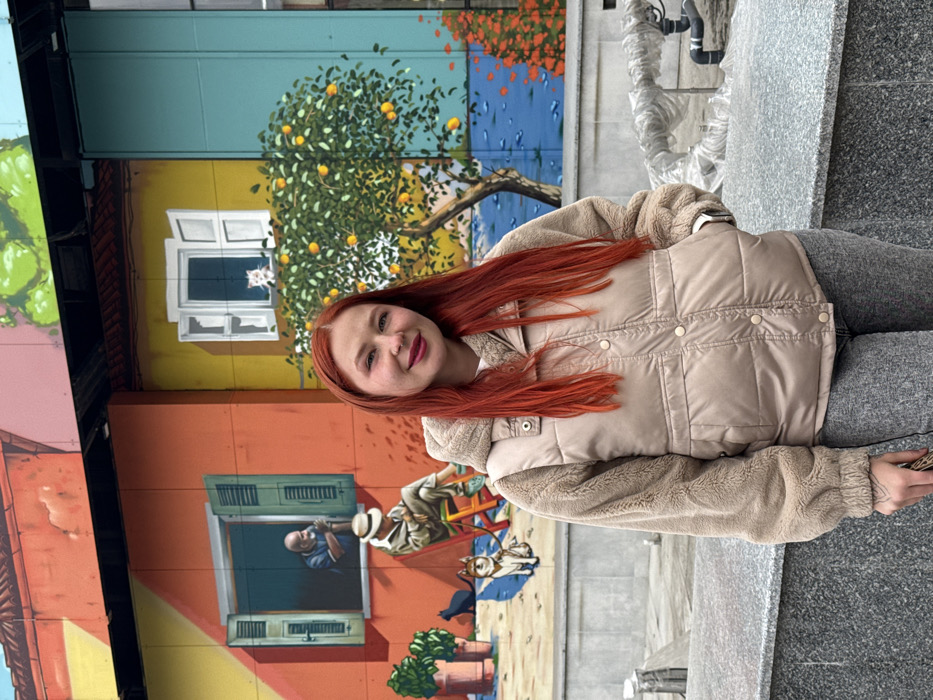 The image depicts a young adult female positioned in front of a multi-panel mural on a building exterior in Rostov-on-Don, Russia. The female, possessing long, straight, bright red hair, a light complexion, and a nostril piercing on her left side, is seen from the waist up. She wears a beige hooded puffer vest over a light brown fleece jacket and grey trousers. Her gaze is directed at the camera, exhibiting a slight smile. Her right hand is in her vest pocket, and her left hand partially grips a dark object out of frame. The vibrant mural behind her portrays a stylized street scene: the left panel features an orange building with an open window revealing an older man looking out, and a seated man with a hat, a small dog, and a black cat below. The central panel displays a yellow building with a white window frame containing a white cat, and a large orange tree with visible fruits. The right panel shows a blue background with orange foliage. In the immediate foreground, a grey granite-like barrier is visible, with clear plastic-wrapped pipes extending from behind it to the right.