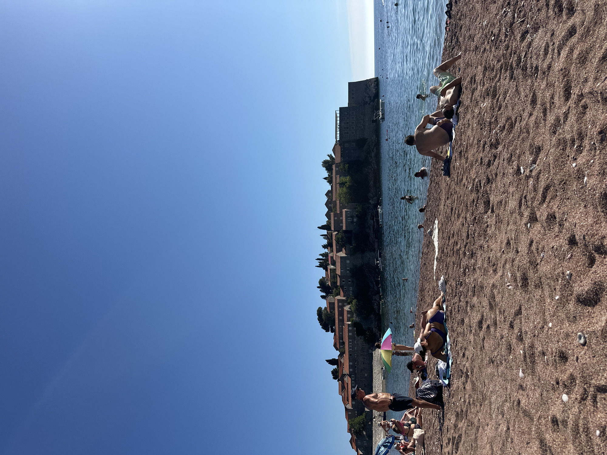 The image depicts a sunny day at a beach in Sveti Stefan, Montenegro, with the historic island of Sveti Stefan in the background. The beach is covered in reddish-brown pebbles. Several people are present on the beach, some sunbathing on towels and others in the water. In the foreground, a man stands on the left side of the frame, and to his right, a woman in a purple bikini is lying on her stomach on a towel. Further to the right, two men are sitting and lying on towels. The water is clear and blue, with several individuals swimming or wading. Boats are visible in the water, and the island itself features stone buildings with red-tiled roofs, nestled among green trees on a rocky outcrop. The sky is clear and bright blue.