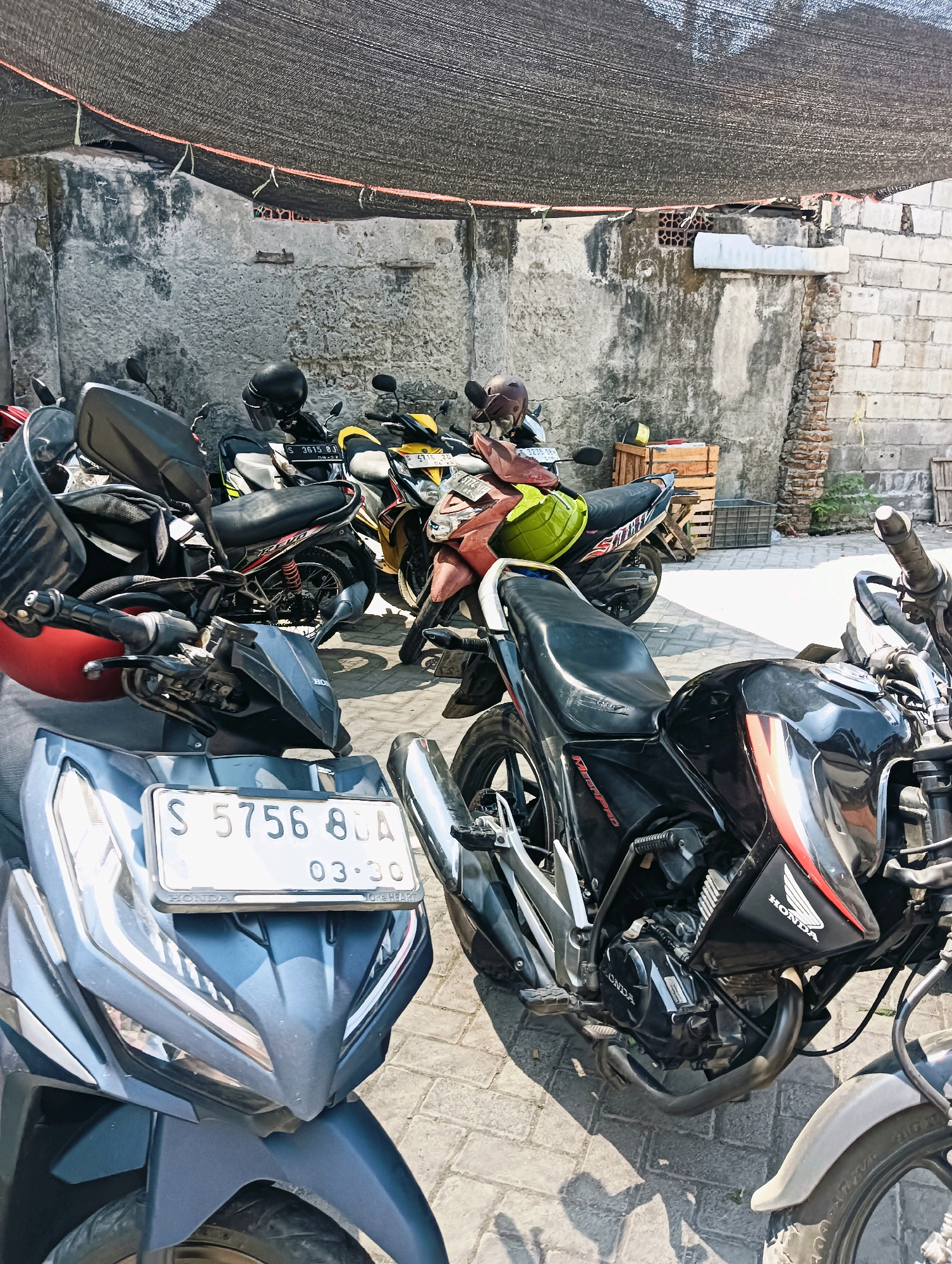 The image depicts a collection of motorcycles parked outdoors under a shaded area, likely a parking lot or an open-air storage space. The background consists of textured, weathered walls and some brickwork, suggesting an urban or semi-urban environment. A dark, mesh-like tarp is stretched overhead, providing shade.
In the foreground, a dark blue scooter with a license plate reading "S 5756 BUA 03.30" is prominently displayed. To its right, a black motorcycle with red and white accents is parked, its engine and exhaust system clearly visible. A Honda logo is visible on its fuel tank. Further back, several other motorcycles are visible, including a yellow and black one with a green helmet on its seat, and a red motorcycle with a brown helmet. Wooden crates and a grey plastic crate are also present in the background. The ground is paved with interlocking bricks.
The scene appears to be static, with no people actively engaged in any activity. The lighting suggests it is daytime, with sunlight casting shadows on the ground. While the location is specified as Losari, Indonesia, there are no specific landmarks or contextual cues within the image itself that definitively confirm this location. The overall impression is that of a typical parking area for motorcycles in a warm climate.
