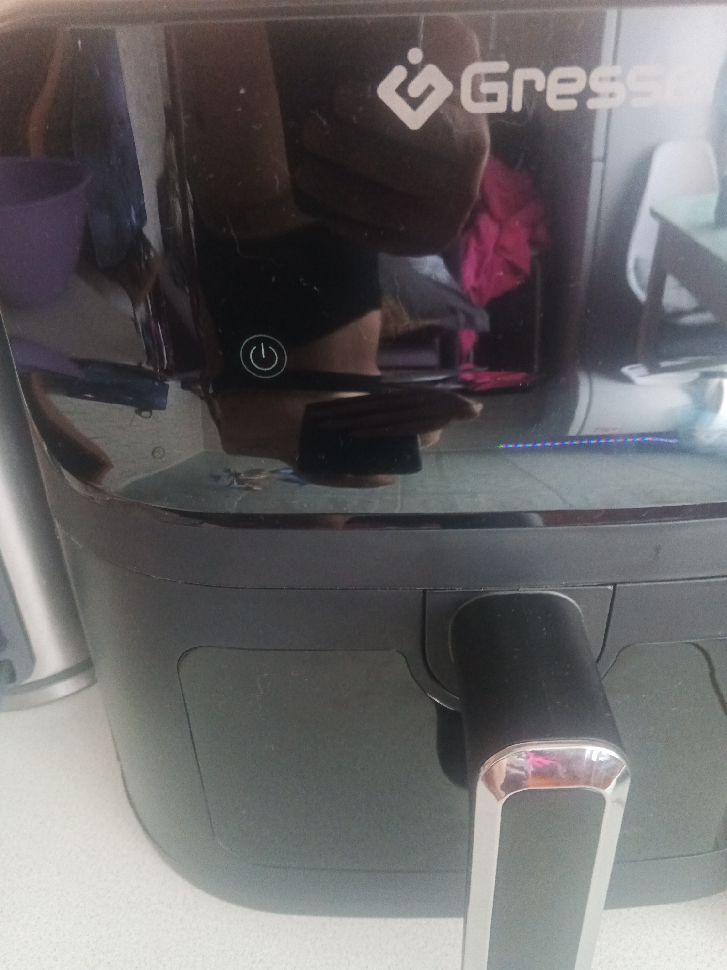 The image shows a close-up, slightly blurry, overhead view of a black air fryer with a silver handle. The air fryer is on a white, speckled countertop. The brand name "Gress" is visible in white lettering on the top surface of the air fryer. A circular power button with a power symbol is also visible. The background is dark and reflective, showing a blurred image of clothing, possibly pink and dark colored, and a white chair. There is a strip of colored lights, possibly blue, red, and green, near the bottom of the air fryer's control panel. No specific location in Biysk, Russia can be determined from the image.
