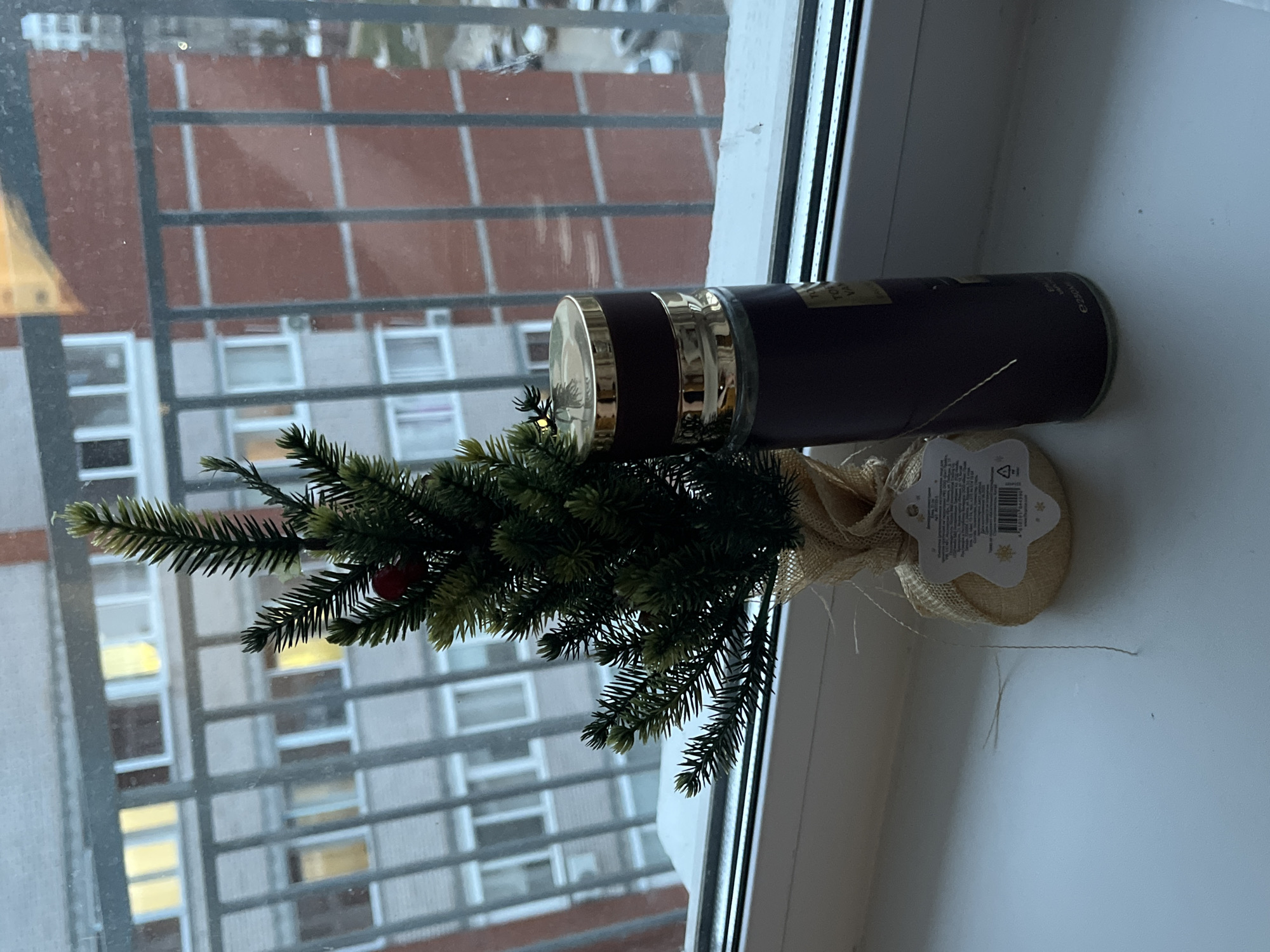 A still life, indoors on a white windowsill in Chaikovsky, Russia. In front of a window with a barred metal frame, an artificial Christmas tree sits in a small burlap sack. A cylindrical, brown bottle with a gold lid lies horizontally behind the tree. The view outside the window features a multi-story building with a repeating pattern of windows.