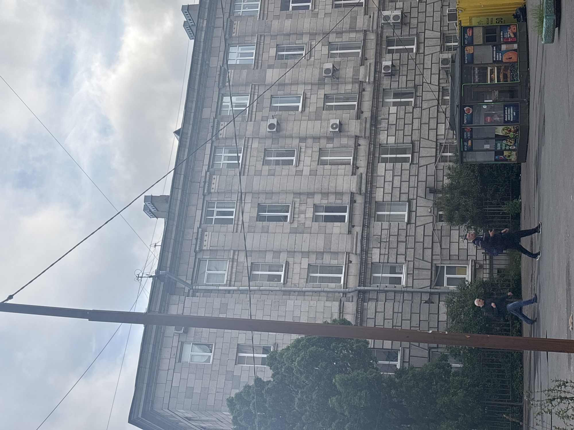 The image depicts an overcast day in Saint Petersburg, Russia, with a multi-story apartment building constructed of grey stone blocks dominating the scene. In the foreground, two men are walking across a street. The man on the left, wearing a dark jacket and blue jeans, walks with his left leg forward. The man on the right, dressed in a dark jacket and black pants, carries a backpack and walks with his right leg forward. To the right of the building, a small kiosk is visible, displaying various food items and advertisements. A yellow shipping container is partially visible behind the kiosk. A prominent brown pole stands vertically in the left mid-ground, crossed by several electrical wires. Lush green trees and bushes line the fence in front of the building, and some vegetation is also present in the lower right corner of the image. The architecture of the building suggests a mid-20th-century Soviet-era design, characterized by its symmetrical facade and repetitive window patterns. The sky is filled with clouds, indicating a typical Saint Petersburg climate.